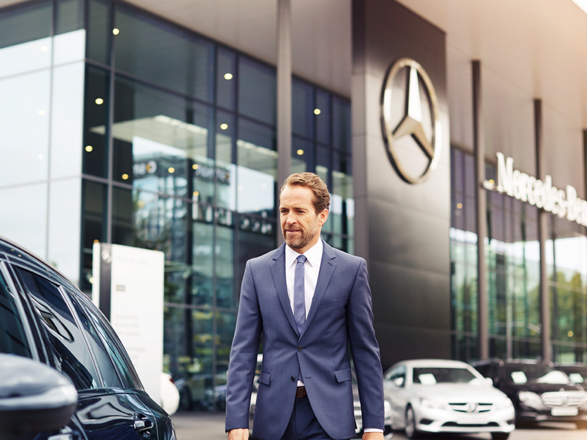 Ein Mann läuft auf sein Auto zu vor einem Mercedes-Benz Kundencenter. Er trägt einen dunklen Anzug.
