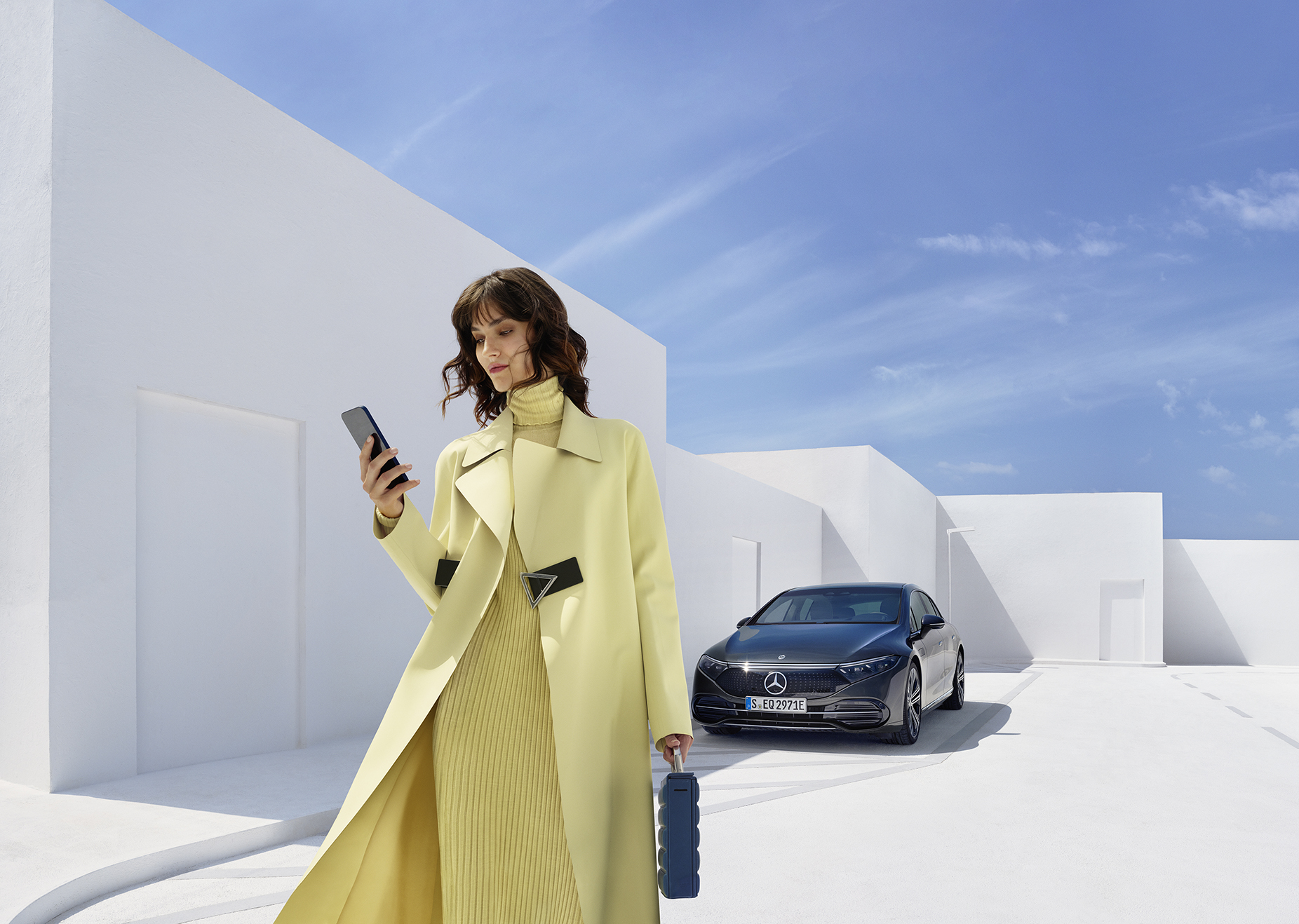 Neuwagen kaufen - so geht's! | Mercedes-Benz Eine Frau in gelbem Outfit schaut auf ihr Handy und steht dabei an einem Straßenrand.