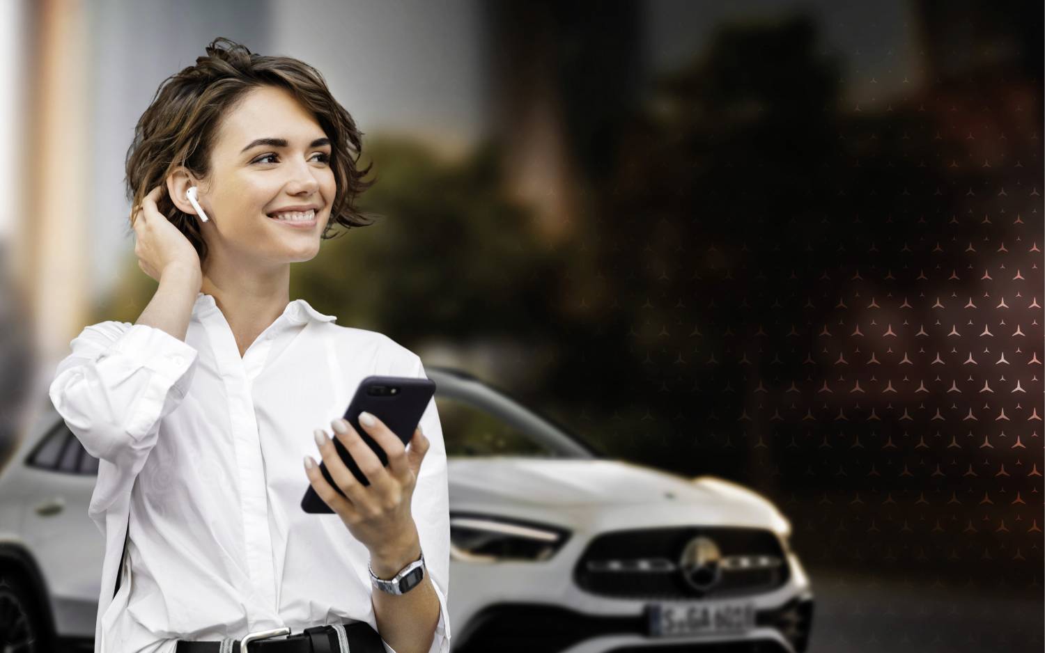 Neuwagen kaufen - so geht's! | Mercedes-Benz Eine Frau mit Bluetooth-Kopfhörer und Handy lächelt in die Ferne.