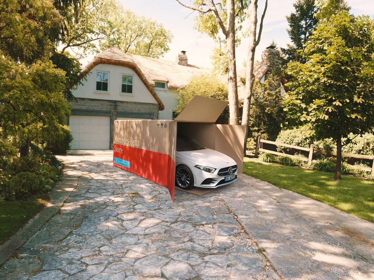 Ein weißes Fahrzeug in einem Pappkarton mit der Aufschrift "Mercedes-Benz Store" steht auf einer Einfahrt.
