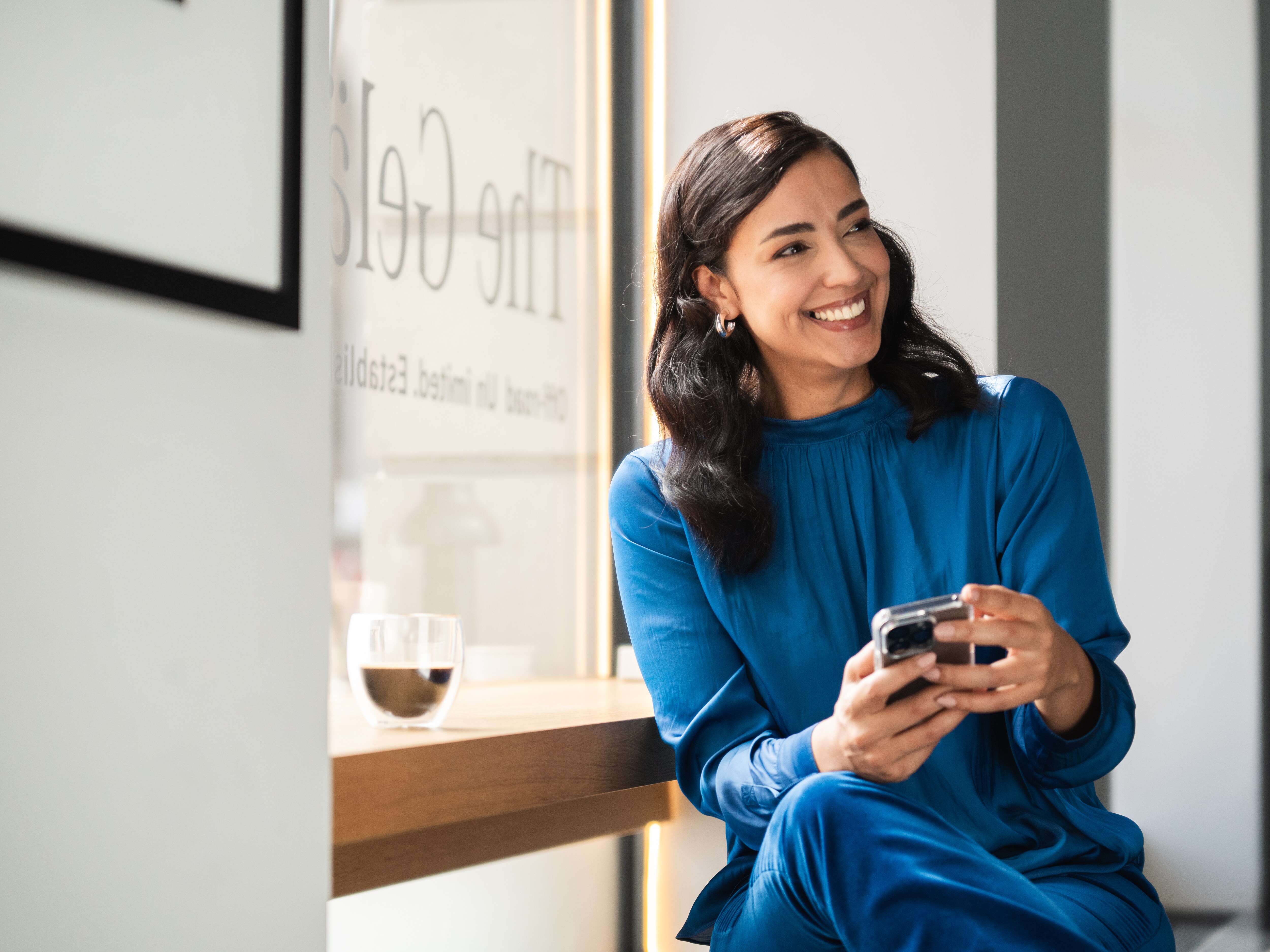 Ein Frau in blauem Kostüm hat ein Telefon in der Hand. Sie schaut lächelnd durch den Raum. 