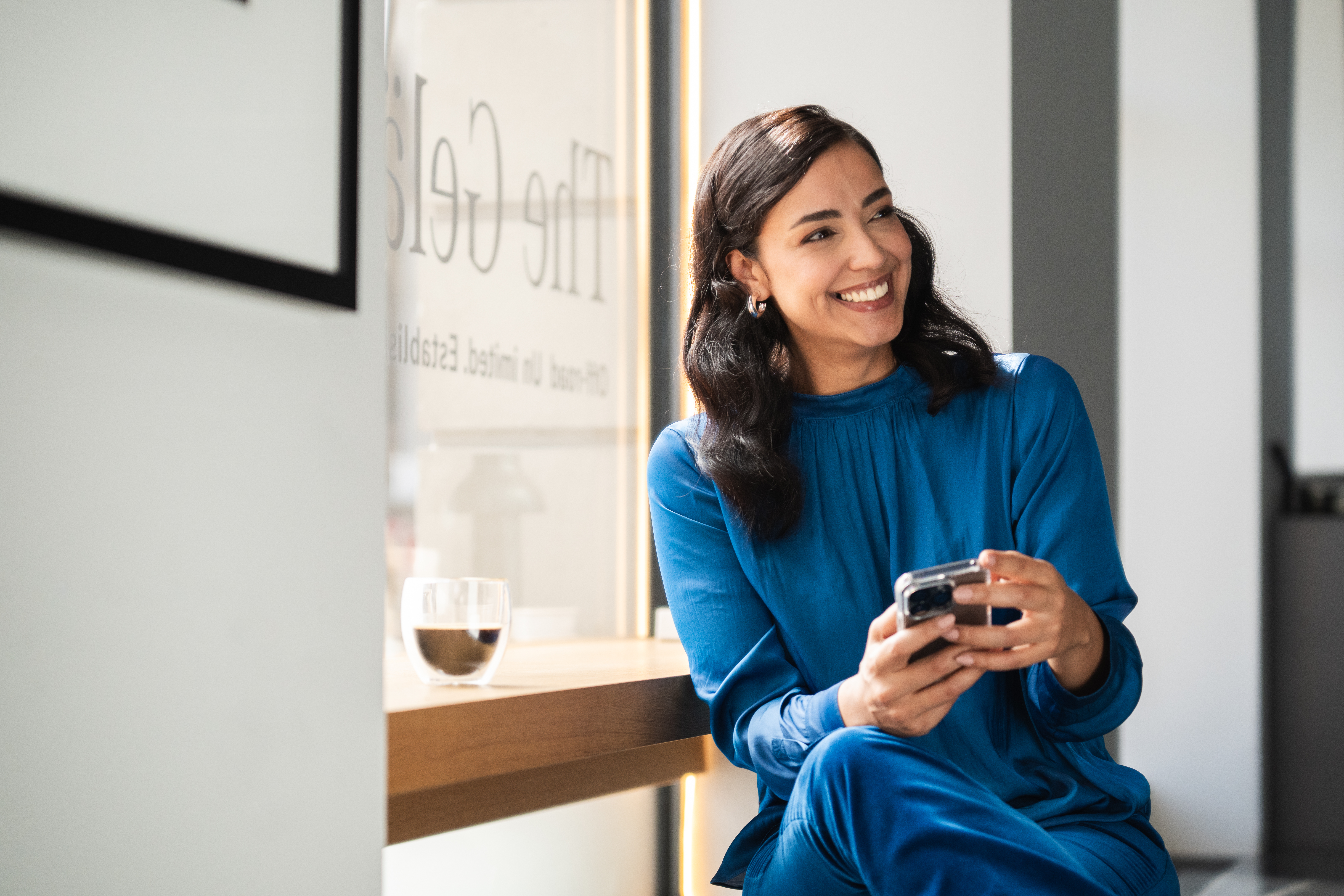 Neuwagen kaufen - so geht's! | Mercedes-Benz Ein Frau in blauem Kostüm hat ein Telefon in der Hand. Sie schaut lächelnd durch den Raum.