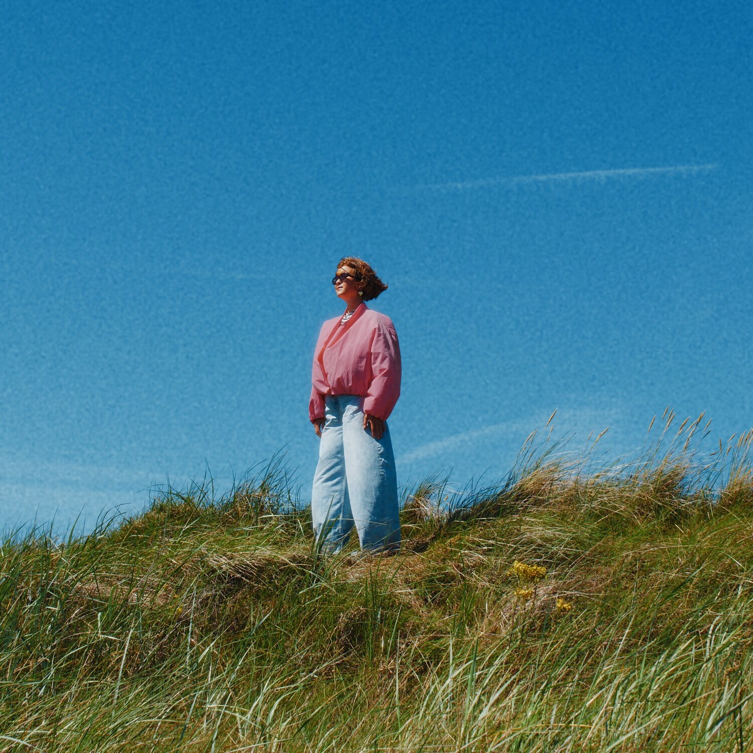 Caro Wahl steht auf einem Grashügel im Wind. Der Himmel ist blau und der Wind kommt ihr entgegen.
