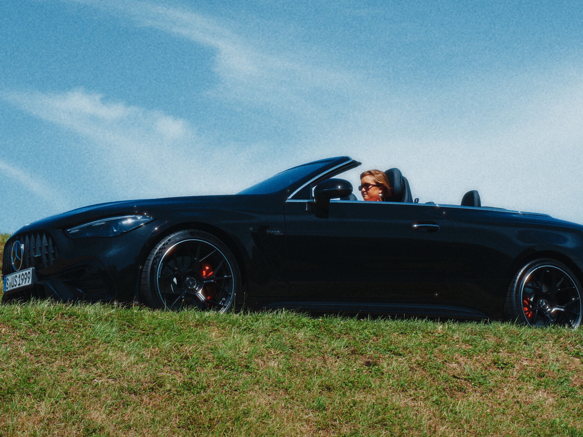 Caro Wahl sitzt in einem schwarzen AMG Carbrio und fährt unter blauem Himmel mit offenem Verdeck.