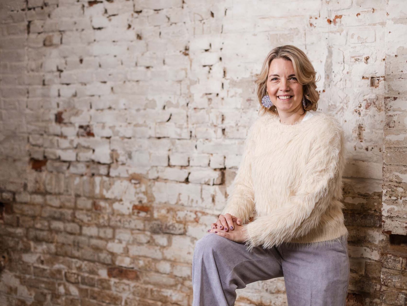 Portrait von Prof. Dr. Christine Laudenbach. Sie lehnt lächelnd an einer Steinmauer und trägt einen hellen Pullover und eine graue Hose.