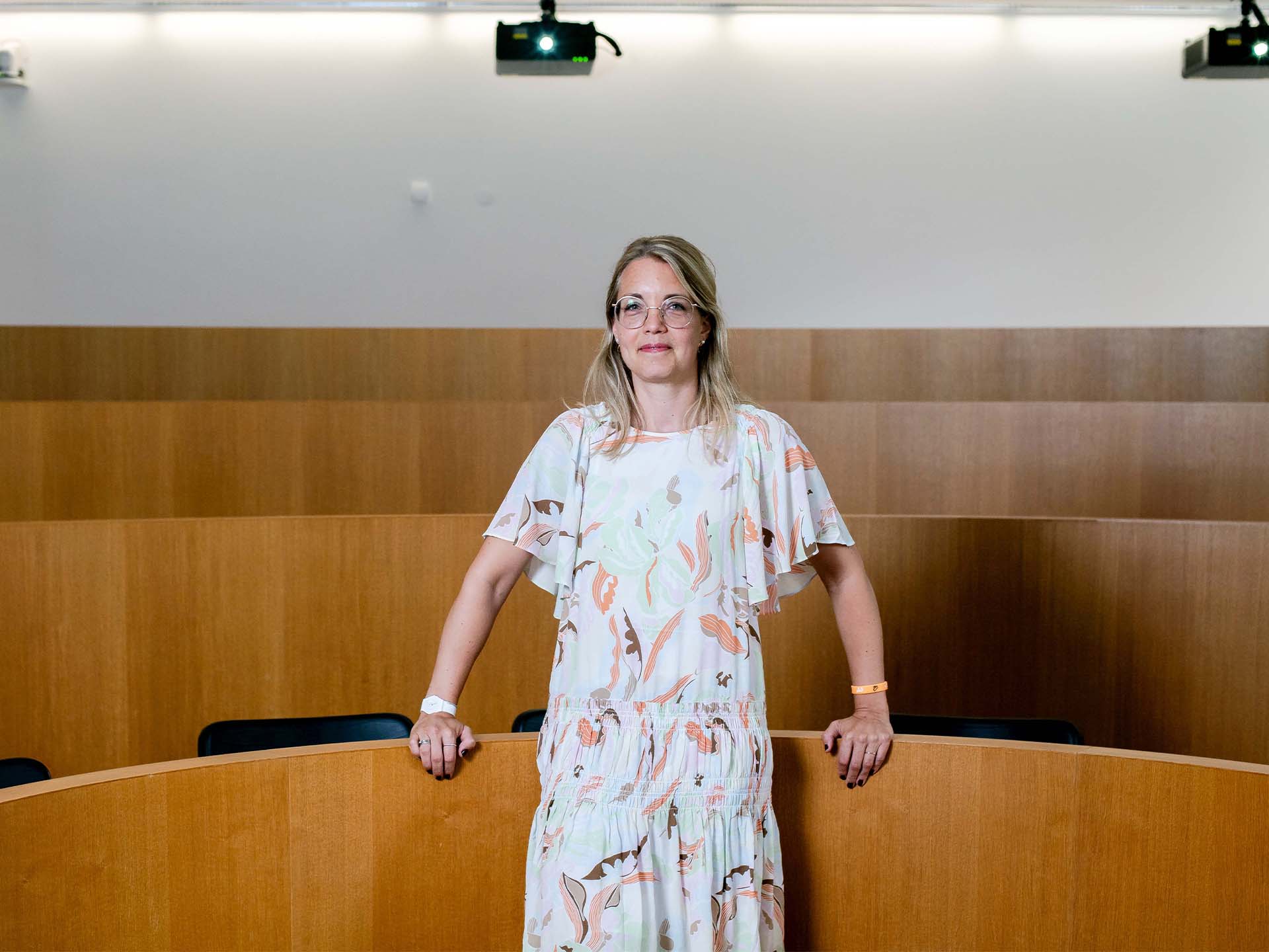 She's Christine Laudenbach | Mercedes-Benz. Prof. Dr. Christine Laudenbach steht in einem hellen Kleid in einem leeren Hörsaal.