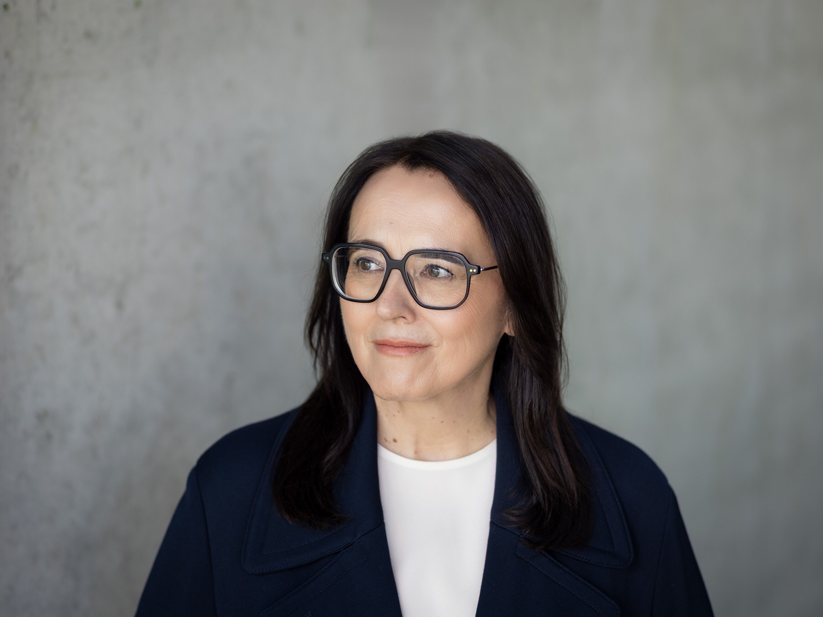 Potrait von Petra Bock vor einer grauen Steinwand. Petra Bock schaut nach links und lächelt.
