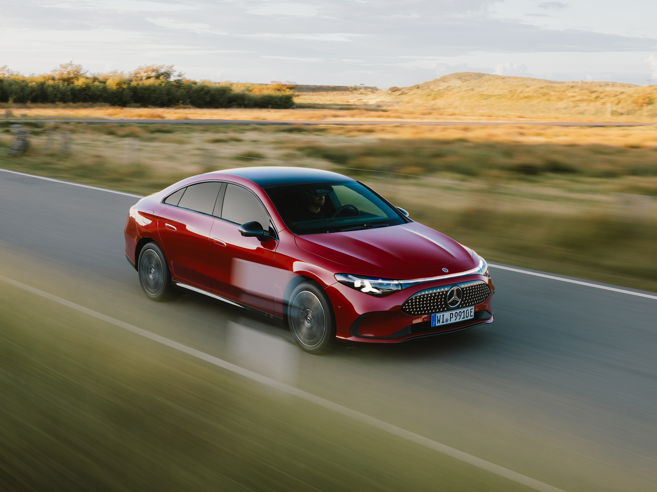Mercedes-Benz x Sylt | Mercedes-Benz. Ein roter elektrischer CLA fährt auf einer Straße zwischen weiten Grasfeldern auf Sylt.