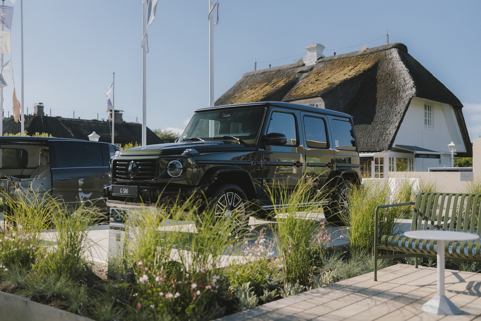 Mercedes-Benz x Sylt | Mercedes-Benz. Eine graue G-Klasse steht auf einer hölzernen Ausstellungsfläche vor einem Haus mit Rieddach.