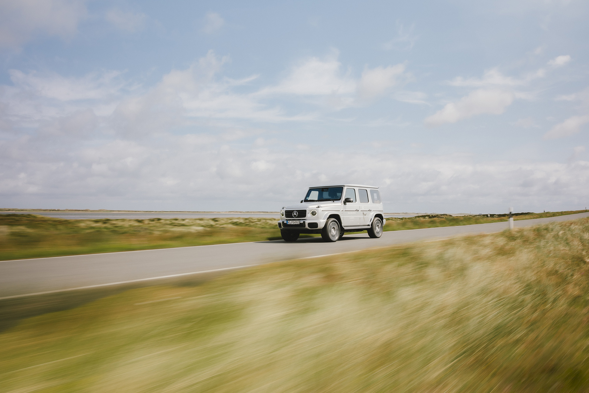 Mercedes-Benz X Sylt | Mercedes-Benz Eine weiße Mercedes-Benz G-Klasse fährt über eine Landstraße am Wasser entlang.