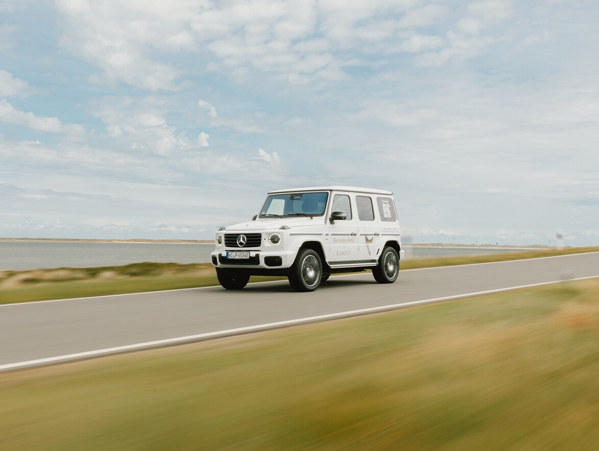 Eine weiße Mercedes-Benz G-Klasse fährt über eine Landstraße am Wasser entlang.