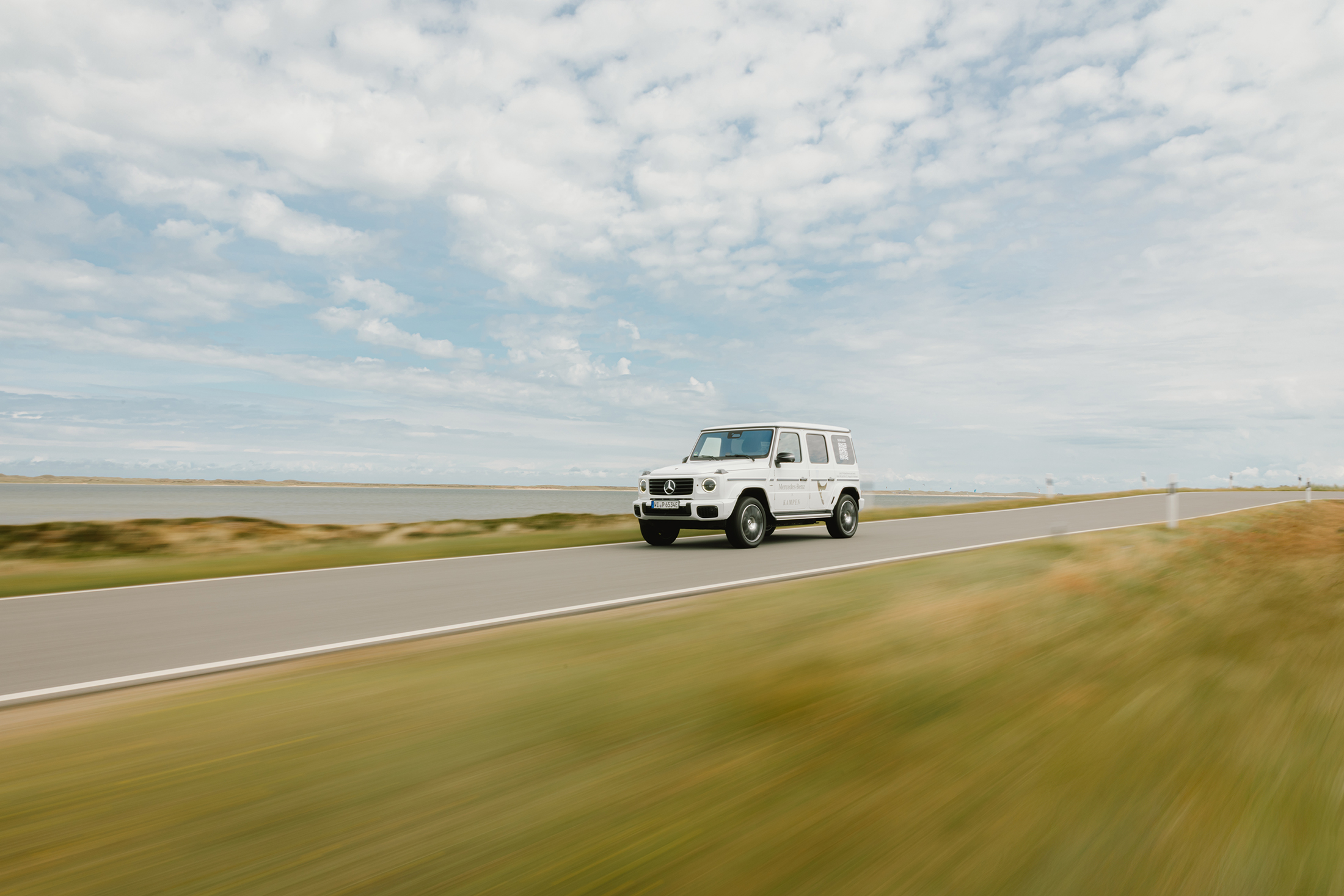 Mercedes-Benz X Sylt | Mercedes-Benz Eine weiße Mercedes-Benz G-Klasse fährt über eine Landstraße am Wasser entlang.