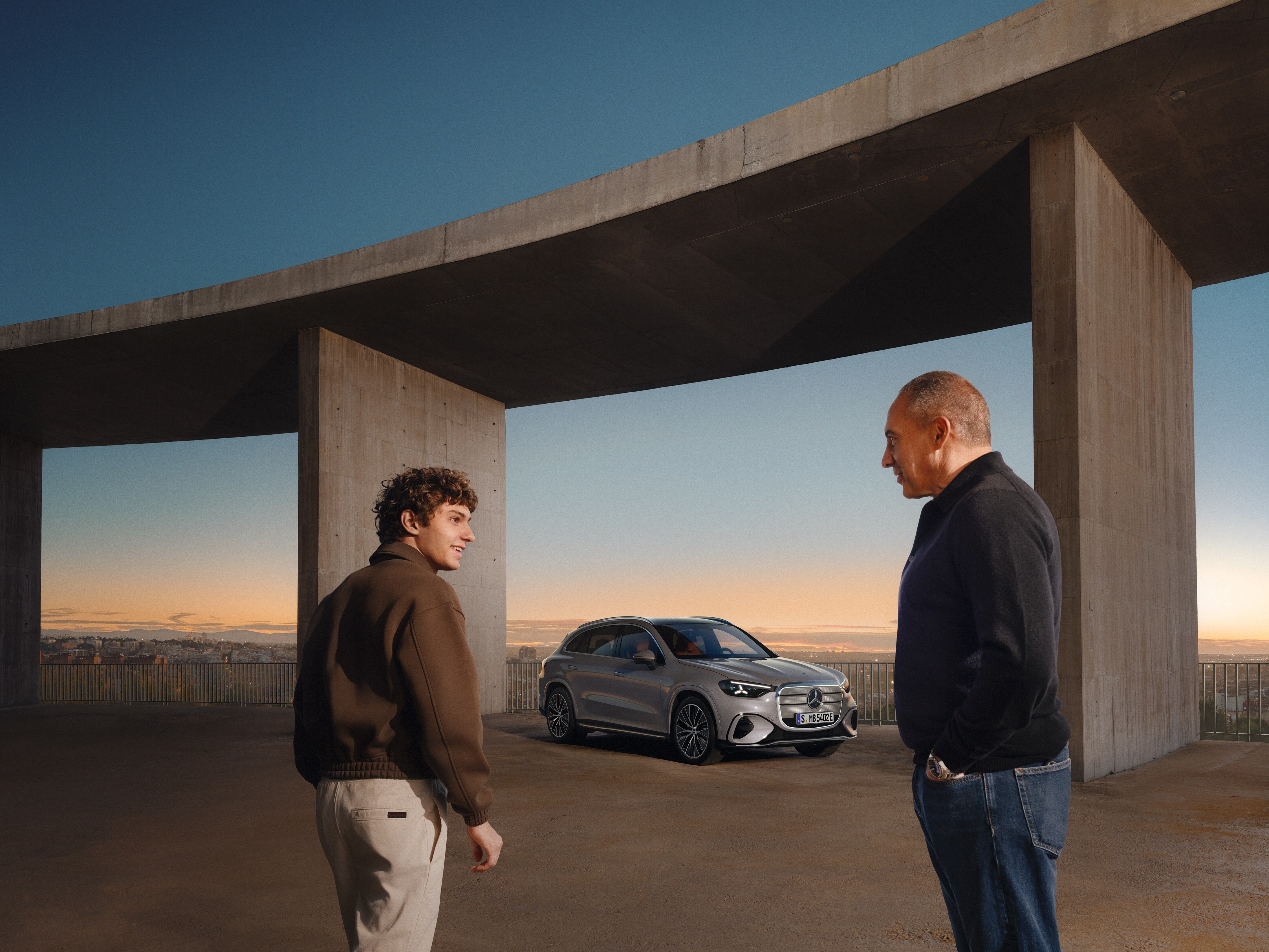 Kimi Antonelli und sein Vater Marco Antonelli stehen vor einer großen Brücke. Unter der Brücke steht der elektrische GLC.