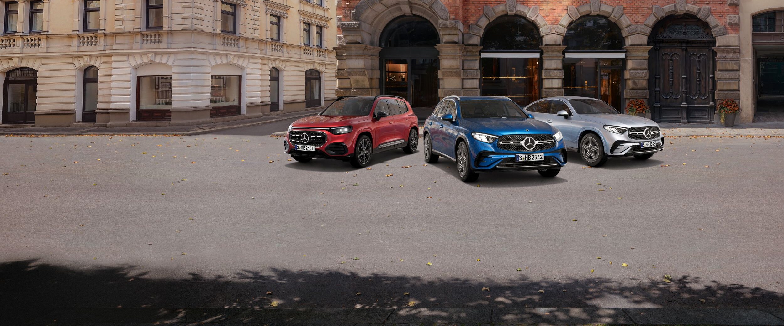  3 Mercedes-Benz Modelle in Rot, Blau und Silber stehen nebeneinander auf einer Straße in einer urbanen Umgebung. Die Sonne scheint.