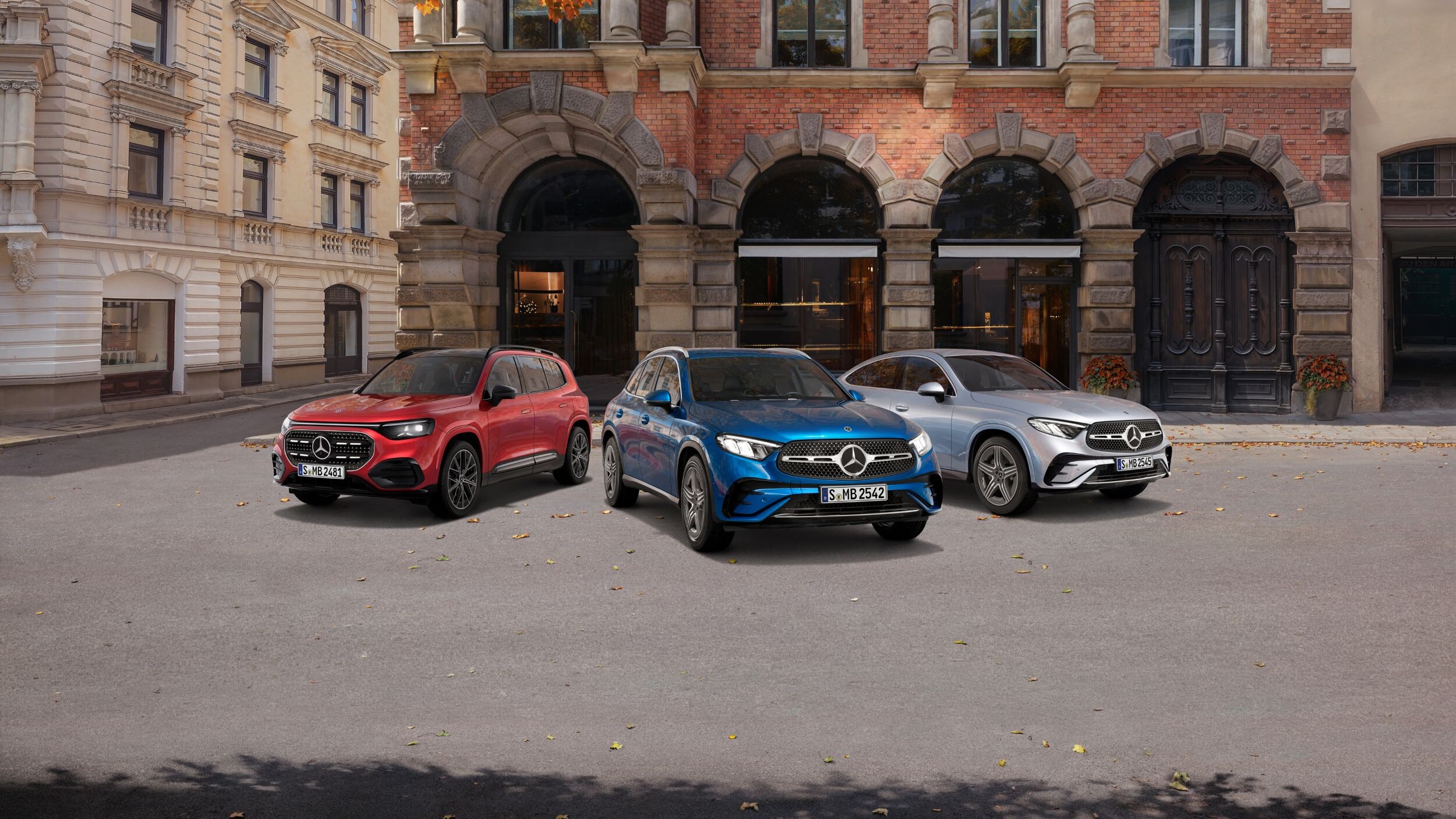 3 Mercedes-Benz Modelle in Rot, Blau und Silber stehen nebeneinander auf einer Straße in einer urbanen Umgebung. Die Sonne scheint.