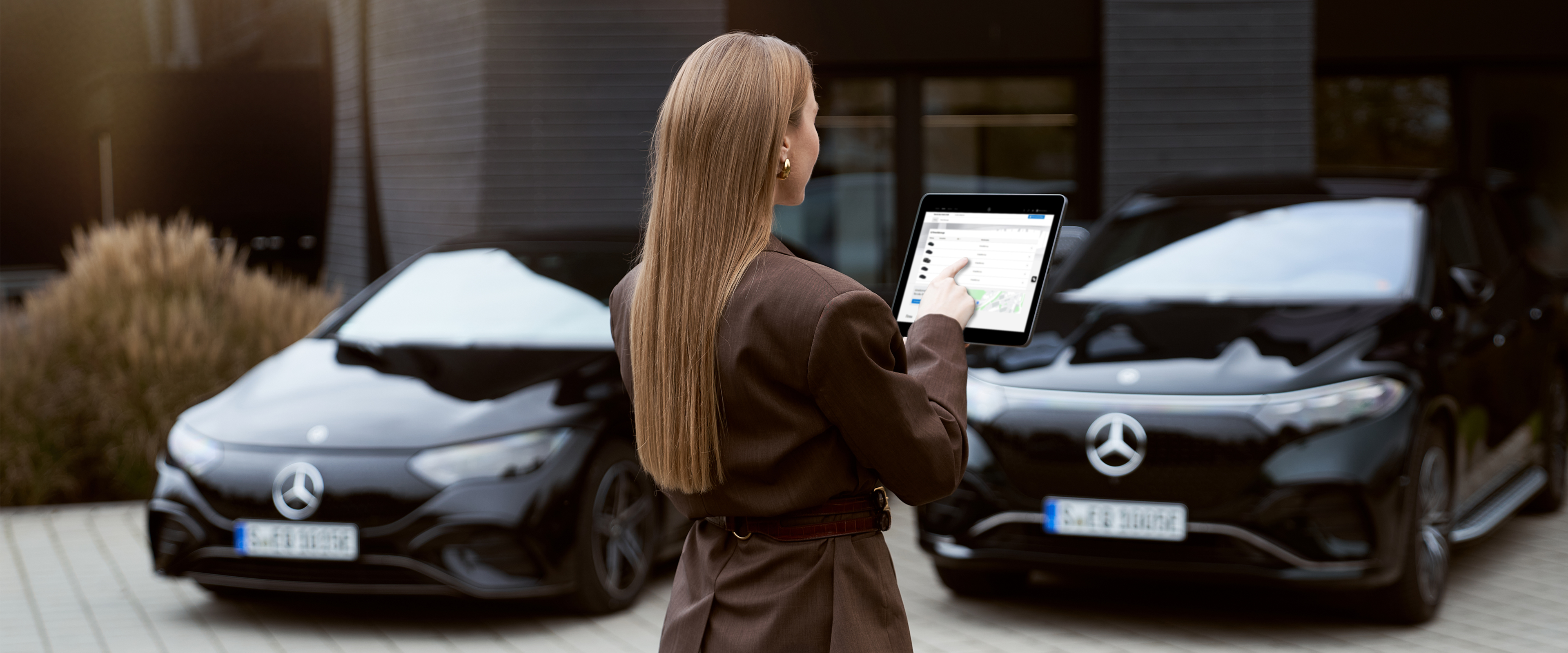 Fuhrpark online konfigurieren & verwalten | Mercedes-Benz Zwei schwarze Mercedes-Benz Fahrzeuge, parkend vor einem Gebäude. Eine Frau steht mit dem Rücken zum Bild und hat ein Tablett in der Hand-