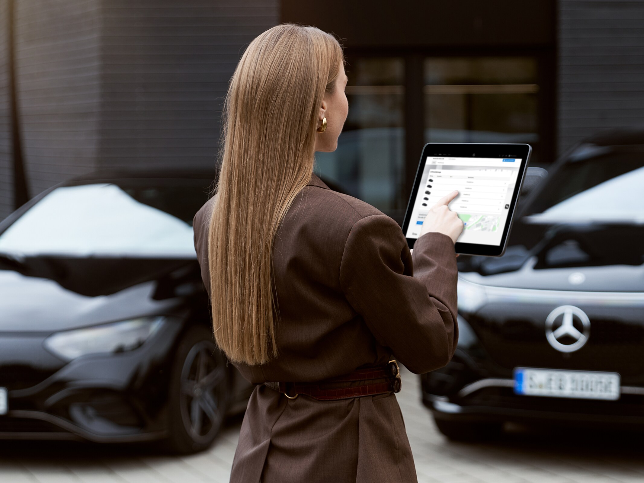 Zwei schwarze Mercedes-Benz Fahrzeuge, parkend vor einem Gebäude. Eine Frau steht mit dem Rücken zum Bild und hat ein Tablett in der Hand-
