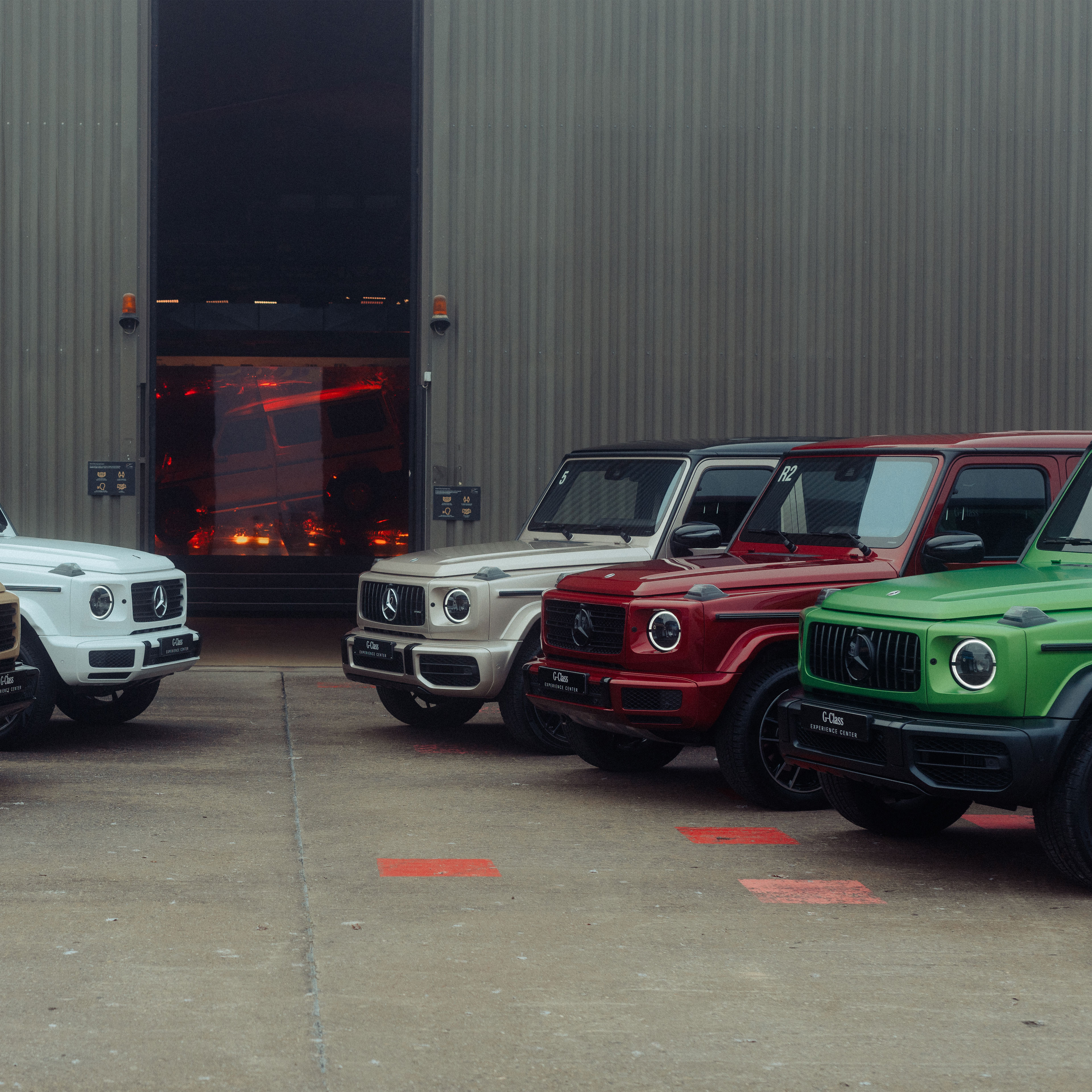 Werksabholung am G-Class Experience Center | Mercedes-Benz. Verschiedene Mercedes-Benz G-Klassen in einer Frontansicht vor dem Werk in Graz.