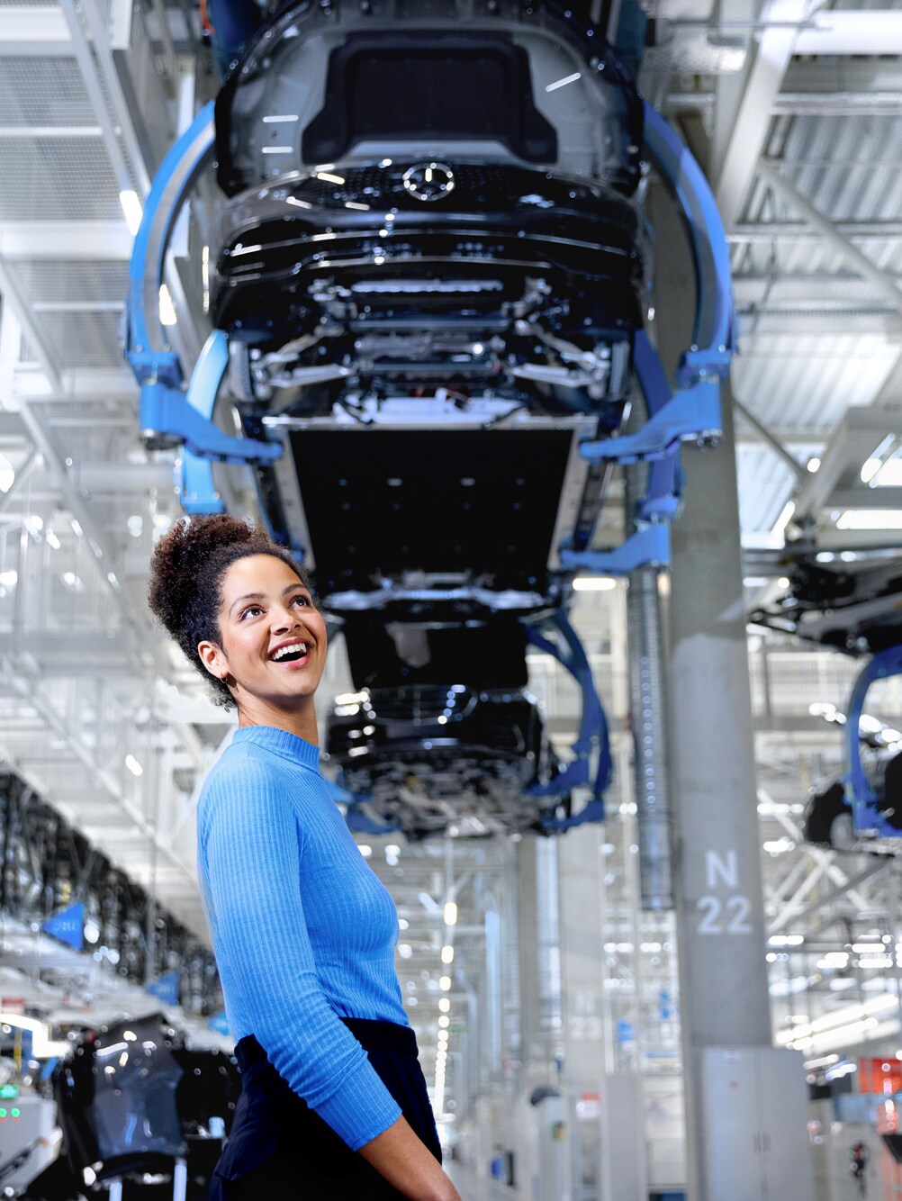 Eine Frau steht lachend neben einem Fahrzeug auf einer Hebebühne in einem Mercedes-Benz Werk.