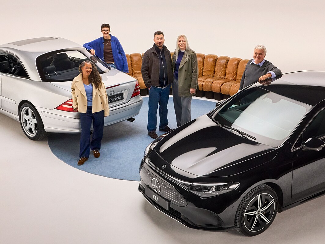 Fünf Personen stehen zwischen einem silbernen und einem schwarzen Mercedes-Benz in einem hell beleuchteten Studio, mit einem beigefarbenen Sofa im Hintergrund.