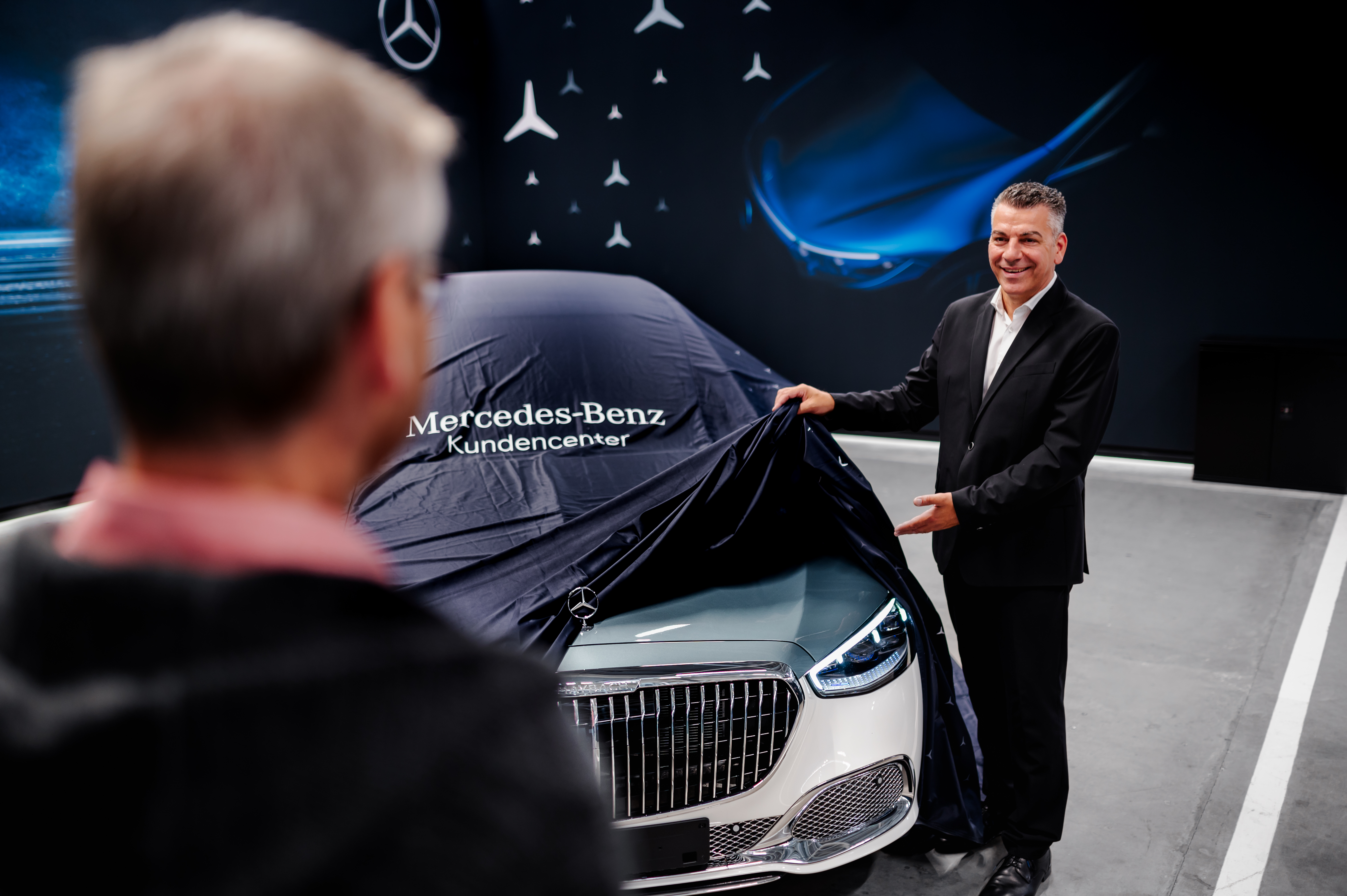 Ihre Fahrzeugabholung | Mercedes-Benz. Zwei Männer stehen an einem Mercedes-Benz Modell, das abgedeckt wird.