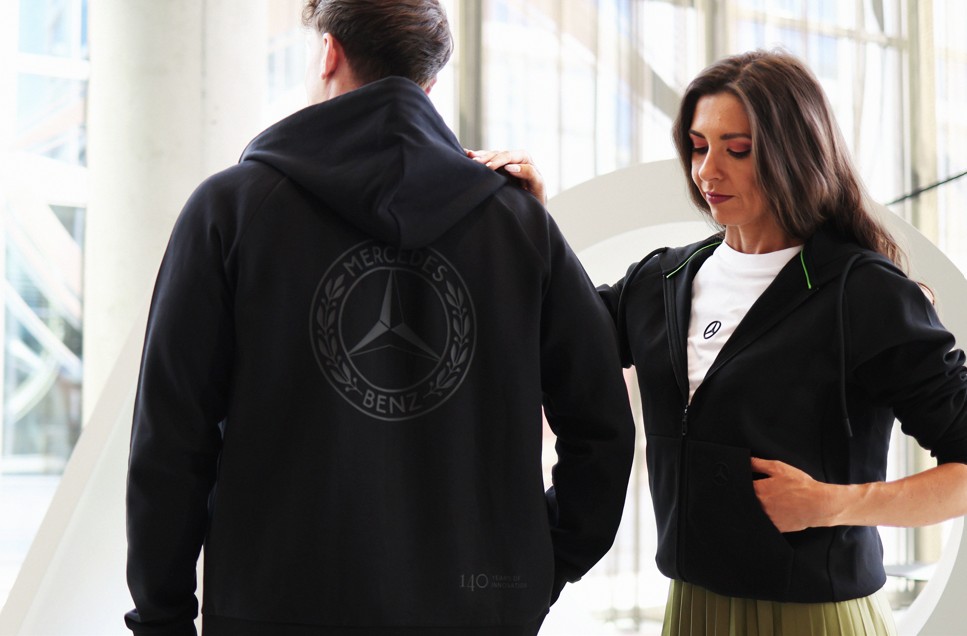Shop im Mercedes‑Benz Kundencenter | Mercedes-Benz. Ein Pärchen in Mercedes Benz Outfit steht in einer Licht durchfluteten Eingangshalle.