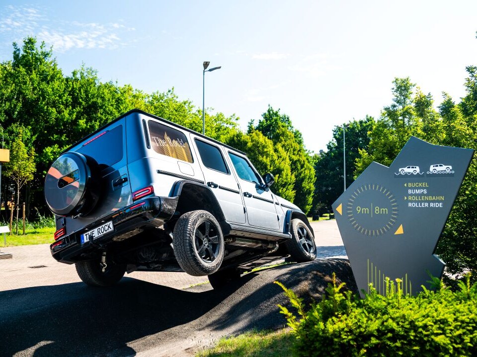Ein silberner Mercedes-Benz G-Klasse fährt über ein unebenes Offroad-Hindernis neben einem Wegweiser für den Geländewagen-Parcours.
