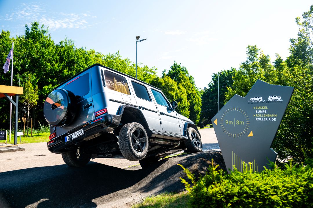 Fahrerlebnisse im Kundencenter | Mercedes-Benz. Ein silberner Mercedes-Benz G-Klasse fährt über ein unebenes Offroad-Hindernis neben einem Wegweiser für den Geländewagen-Parcours.