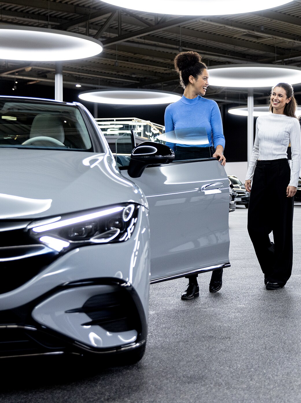 Eine Frau öffnet einem Paar lächelnd die Autotür ihres neuen, silbernen Mercedes-Benz.