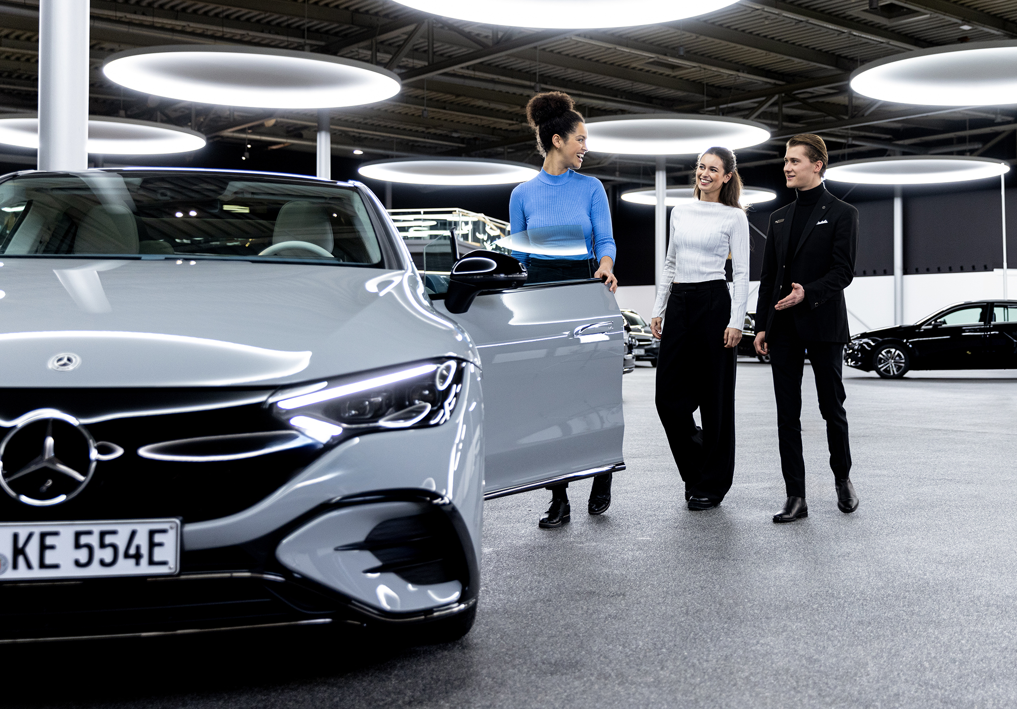 Ihre Fahrzeugabholung | Mercedes-Benz. Eine Frau öffnet einem Paar lächelnd die Autotür ihres neuen, silbernen Mercedes-Benz.