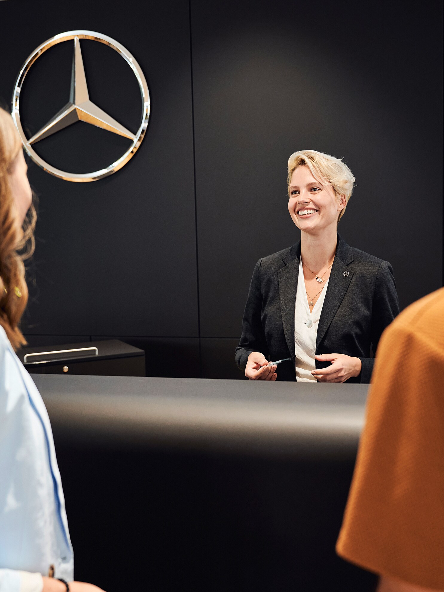 Eine Frau begrüßt eine Frau und einen Mann am Empfang. Dabei lächelt sie beide an.