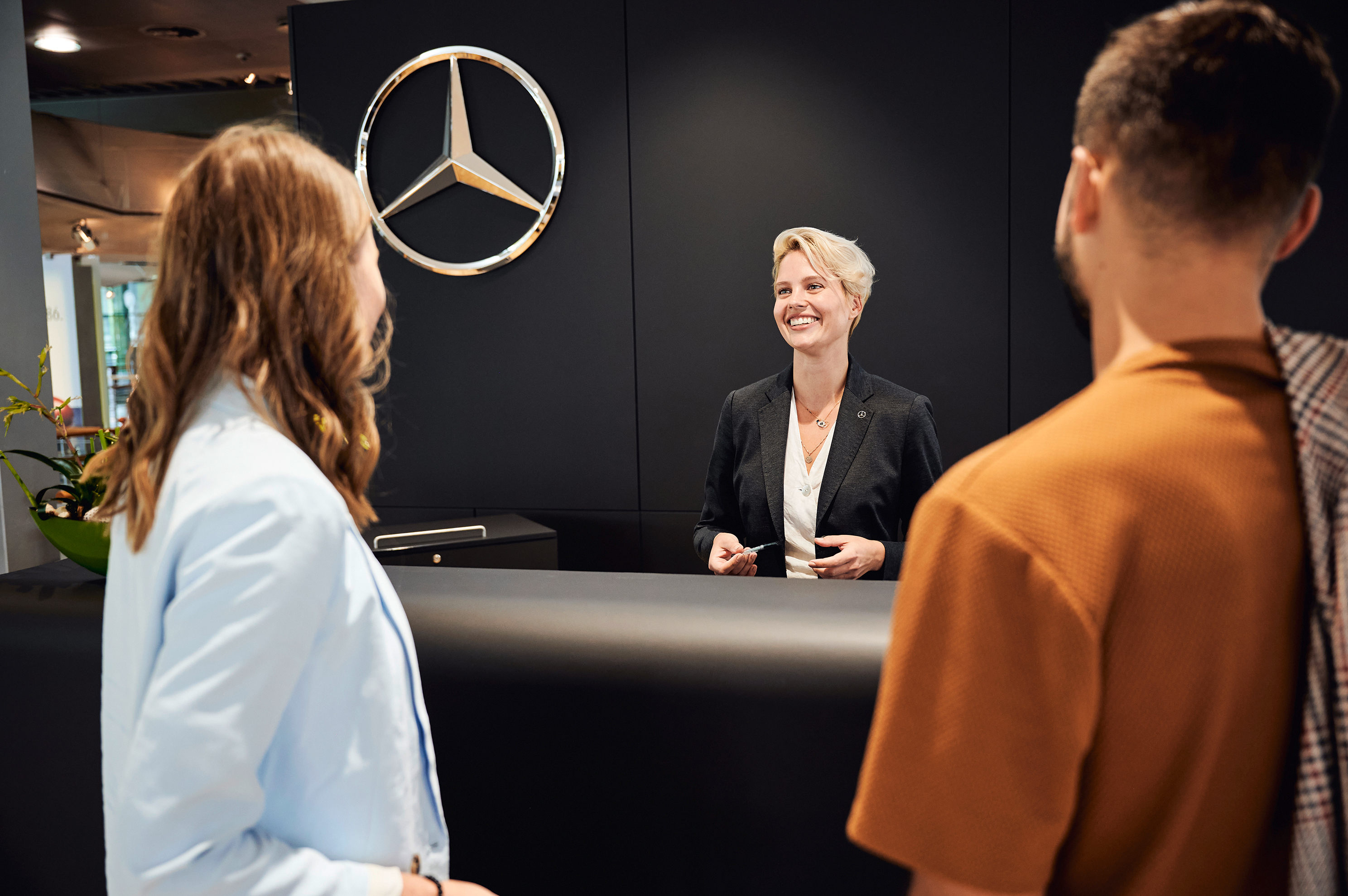 Ihre Fahrzeugabholung | Mercedes-Benz. Eine Frau begrüßt eine Frau und einen Mann am Empfang. Dabei lächelt sie beide an.