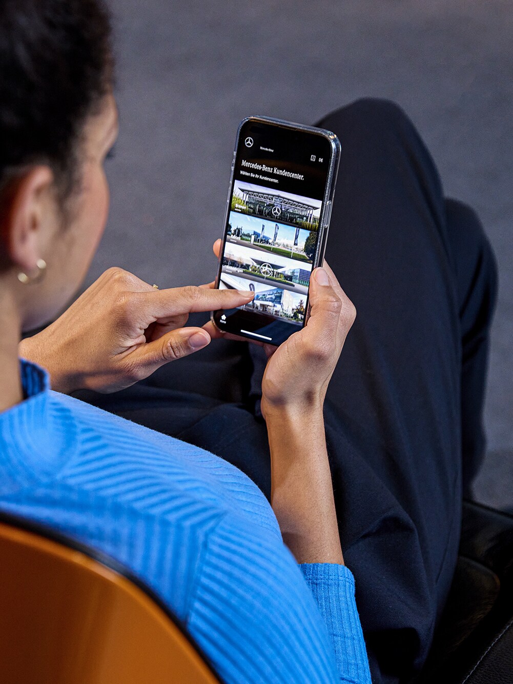 Eine Frau sitzt auf einem Stuhl und schaut auf ihr Telefon. Auf diesem ist die Mercedes-Benz App geöffnet.