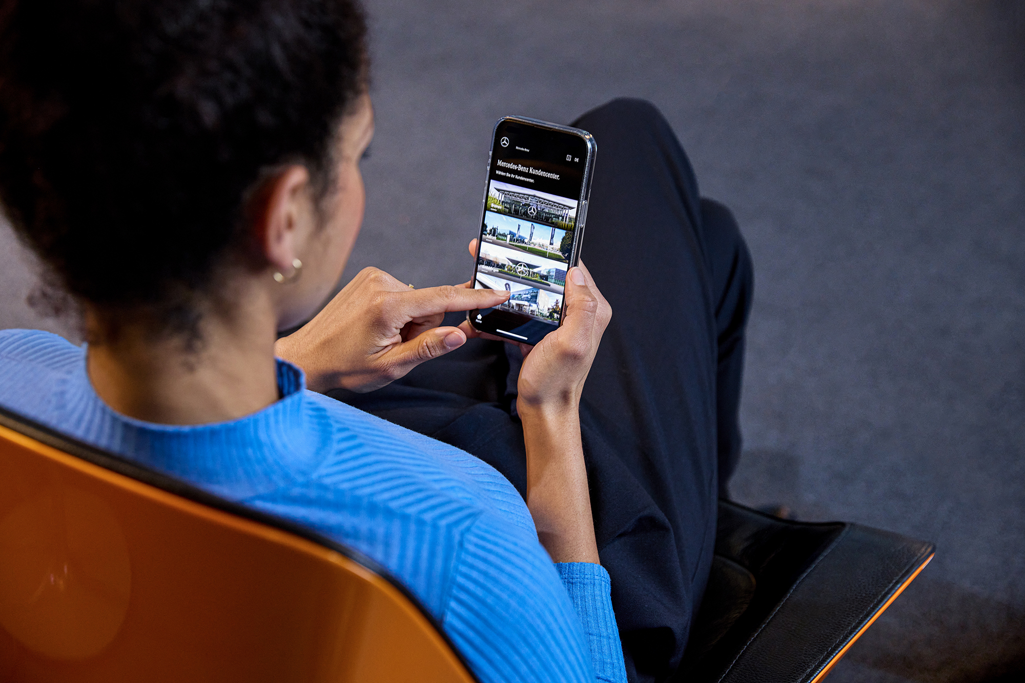 Ihre Fahrzeugabholung | Mercedes-Benz. Eine Frau sitzt auf einem Stuhl und schaut auf ihr Telefon. Auf diesem ist die Mercedes-Benz App geöffnet.