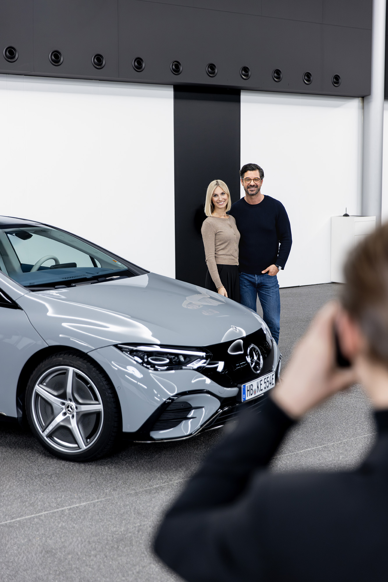 Ihre Fahrzeugabholung | Mercedes-Benz. Ein Paar steht hinter einem hellen Mercedes-Benz Modell und wird dabei fotografiert.