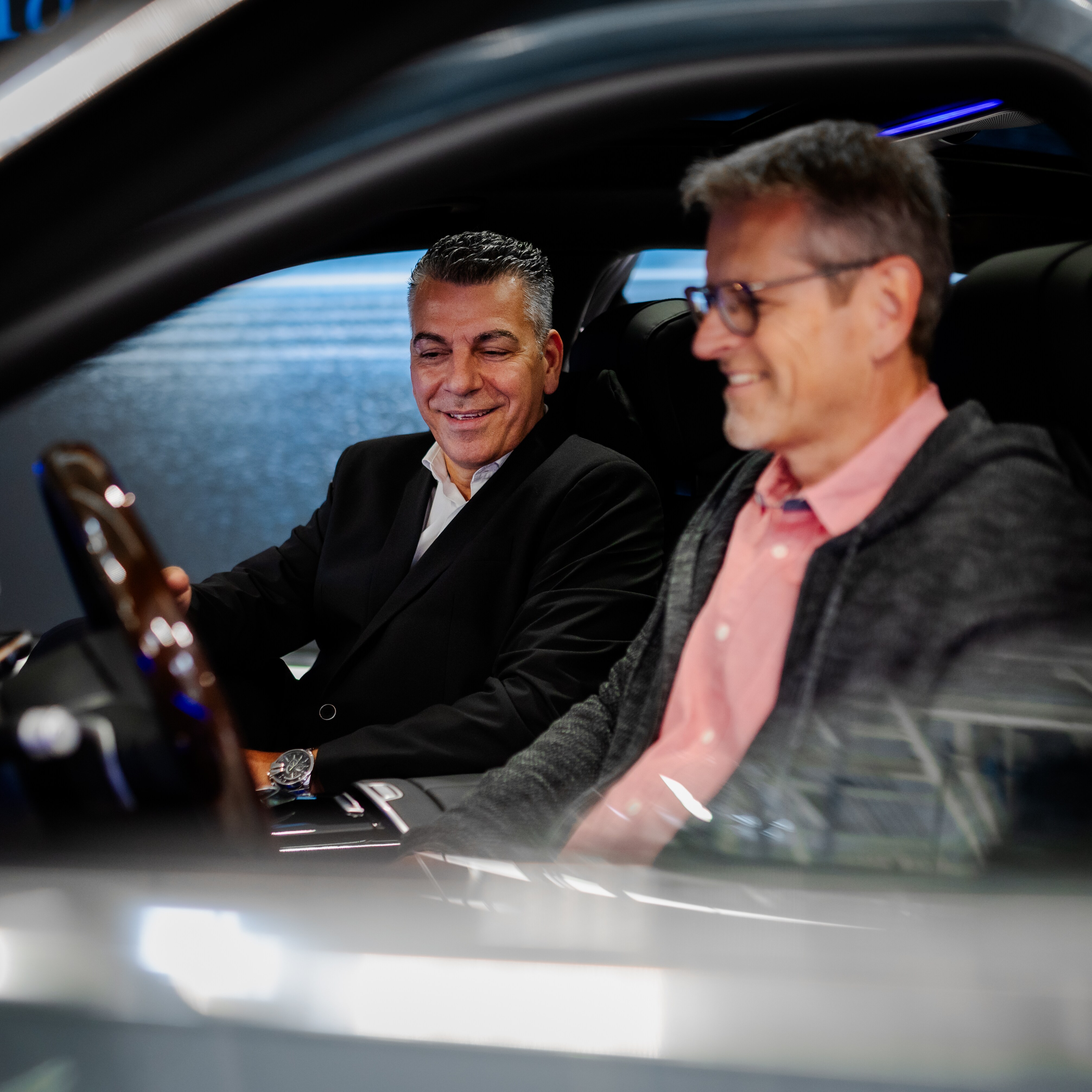 Zwei Männer sitzen in einem Mercedes-Benz und lachen. Dabei erklärt einer dem anderen die Technik des Fahrzeugs.