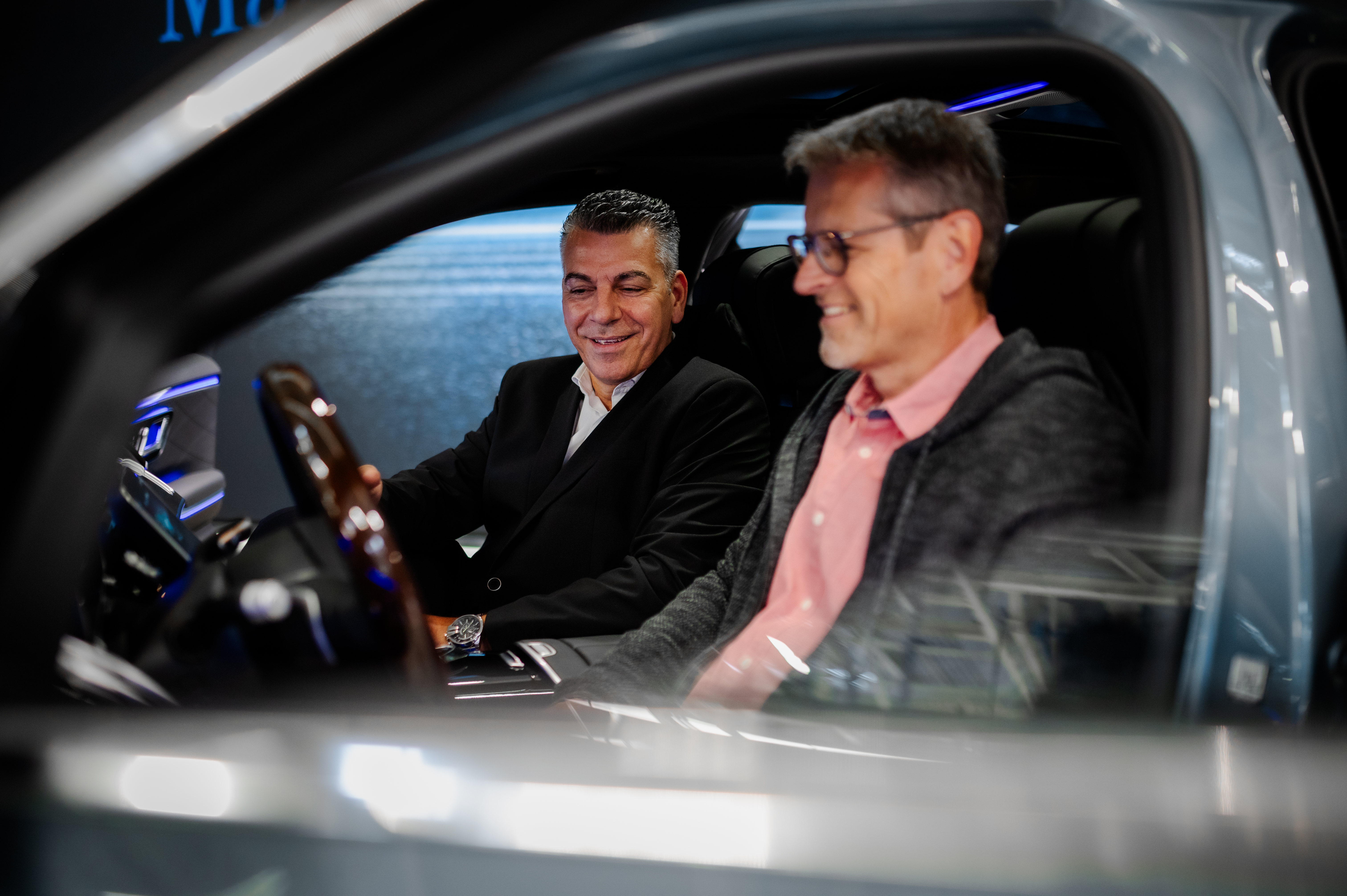 Ihre Fahrzeugabholung | Mercedes-Benz. Zwei Männer sitzen in einem Mercedes-Benz und lachen. Dabei erklärt einer dem anderen die Technik des Fahrzeugs.