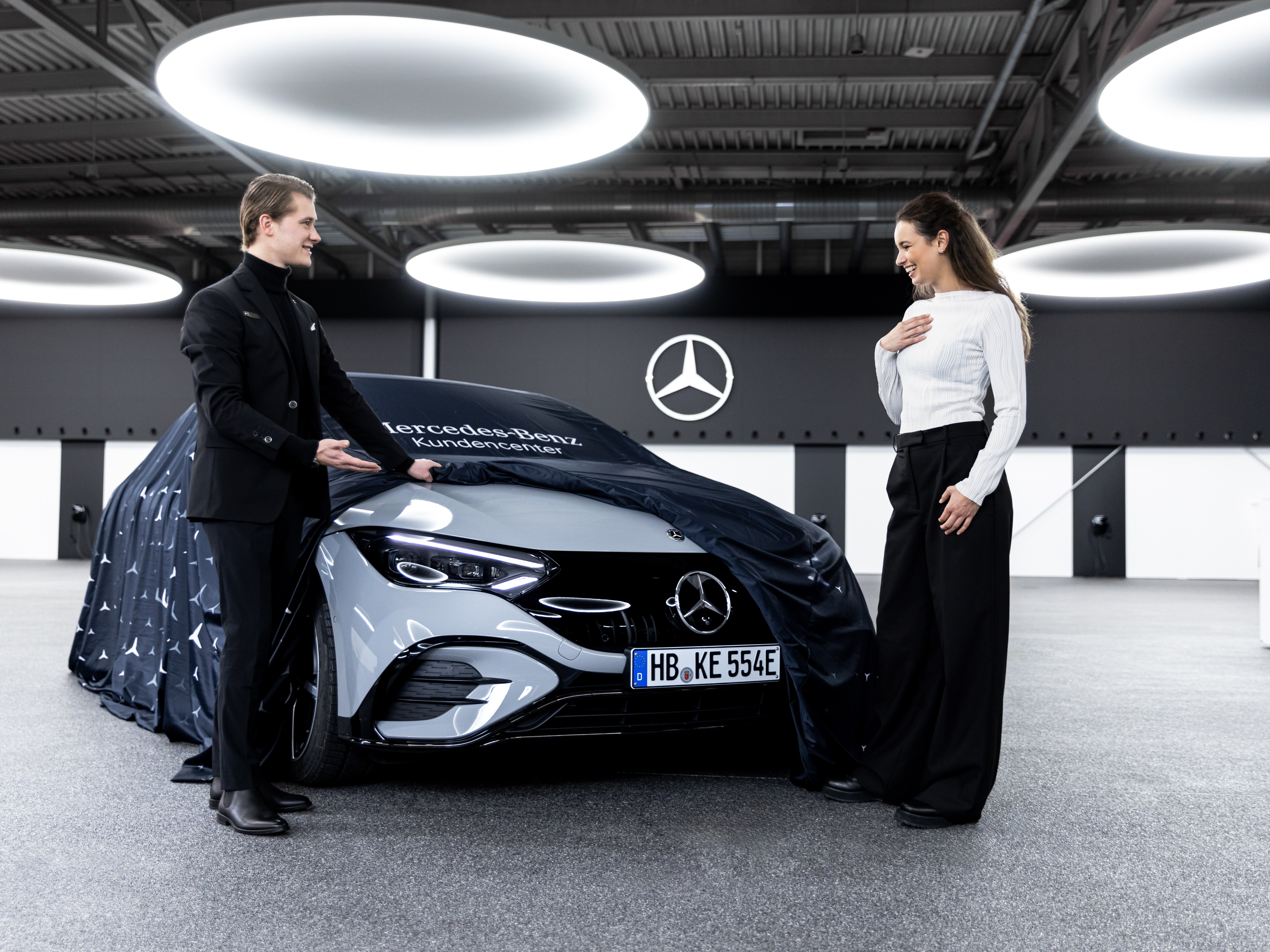 Ein Mann präsentiert einer Frau einen neuen Mercedes-Benz. Die Frau ist begeistert und er lächelt.