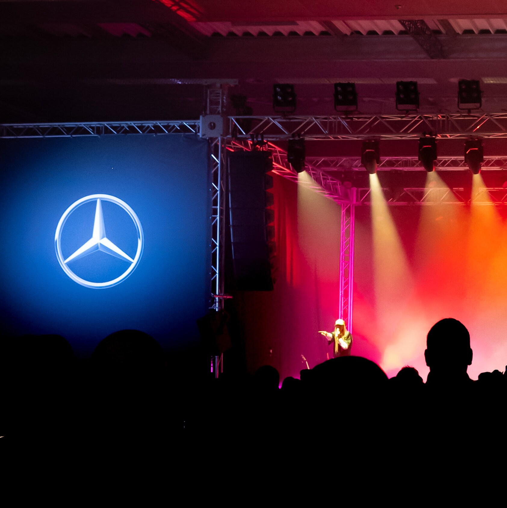 Eine rot beleuchtete Bühne mit seitlich leuchtenden Mercedes-Sternen vor einer Menschenmenge. 