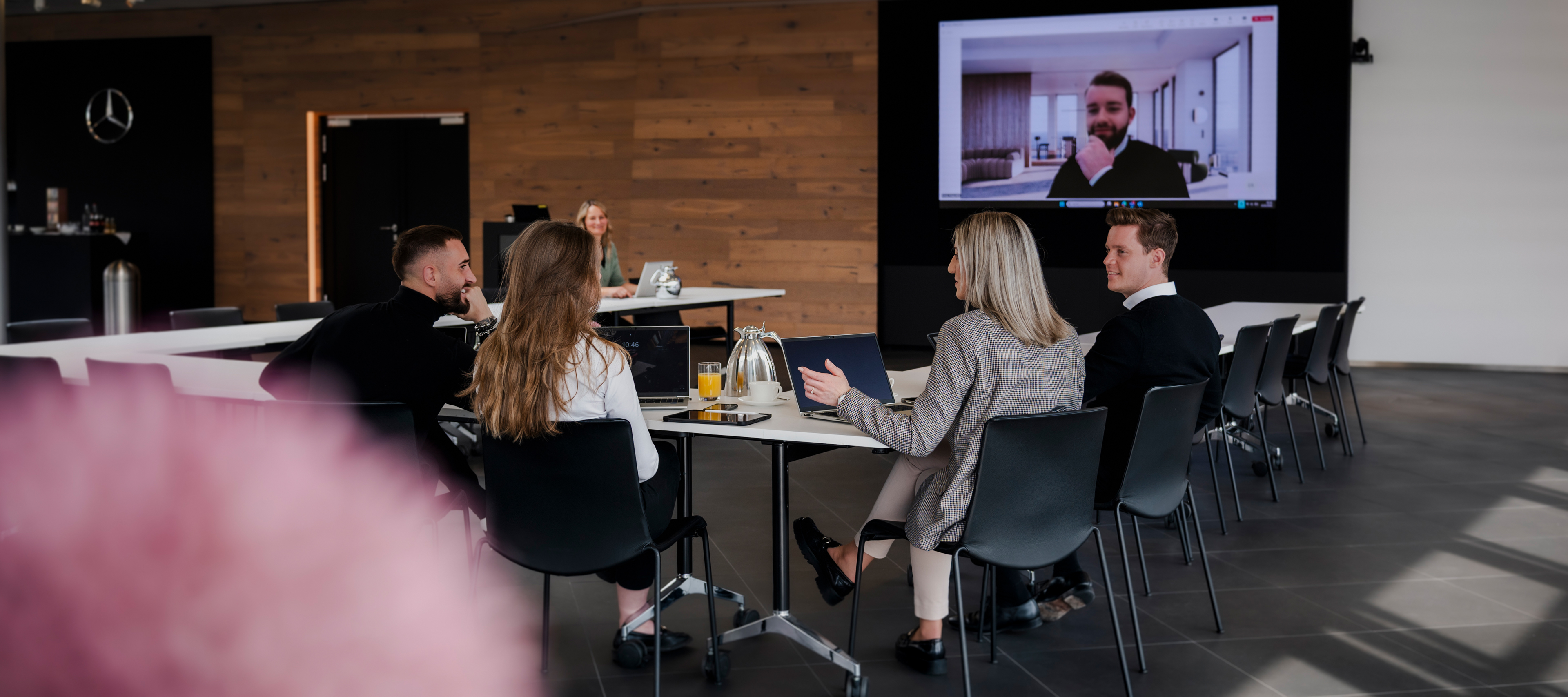 Konferenzen im Kundencenter | Mercedes‑Benz. Fünf Personen sitzen an einem Konferenztisch und schauen auf einen Videoleinwand mit einem weiteren Gesprächsteilnehmer.