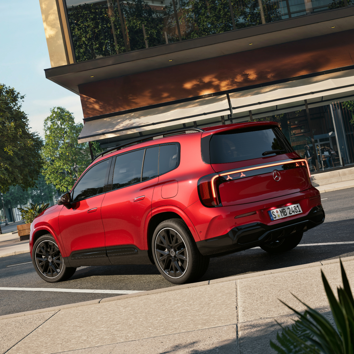 Warum entscheiden | Mercedes-Benz Ein roter GLB SUV steht auf einem sonnigen Parkplatz vor einem braunen Gebäude.