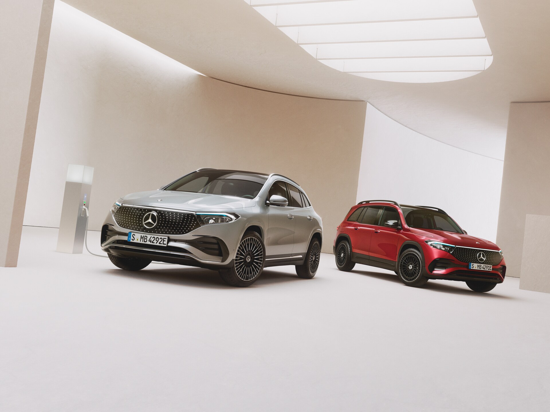 Ein roter Mercedes-Benz EQB und ein grauer Mercedes-Benz EQA stehen auf einem hellen Untergrund an einer Ladesäule.