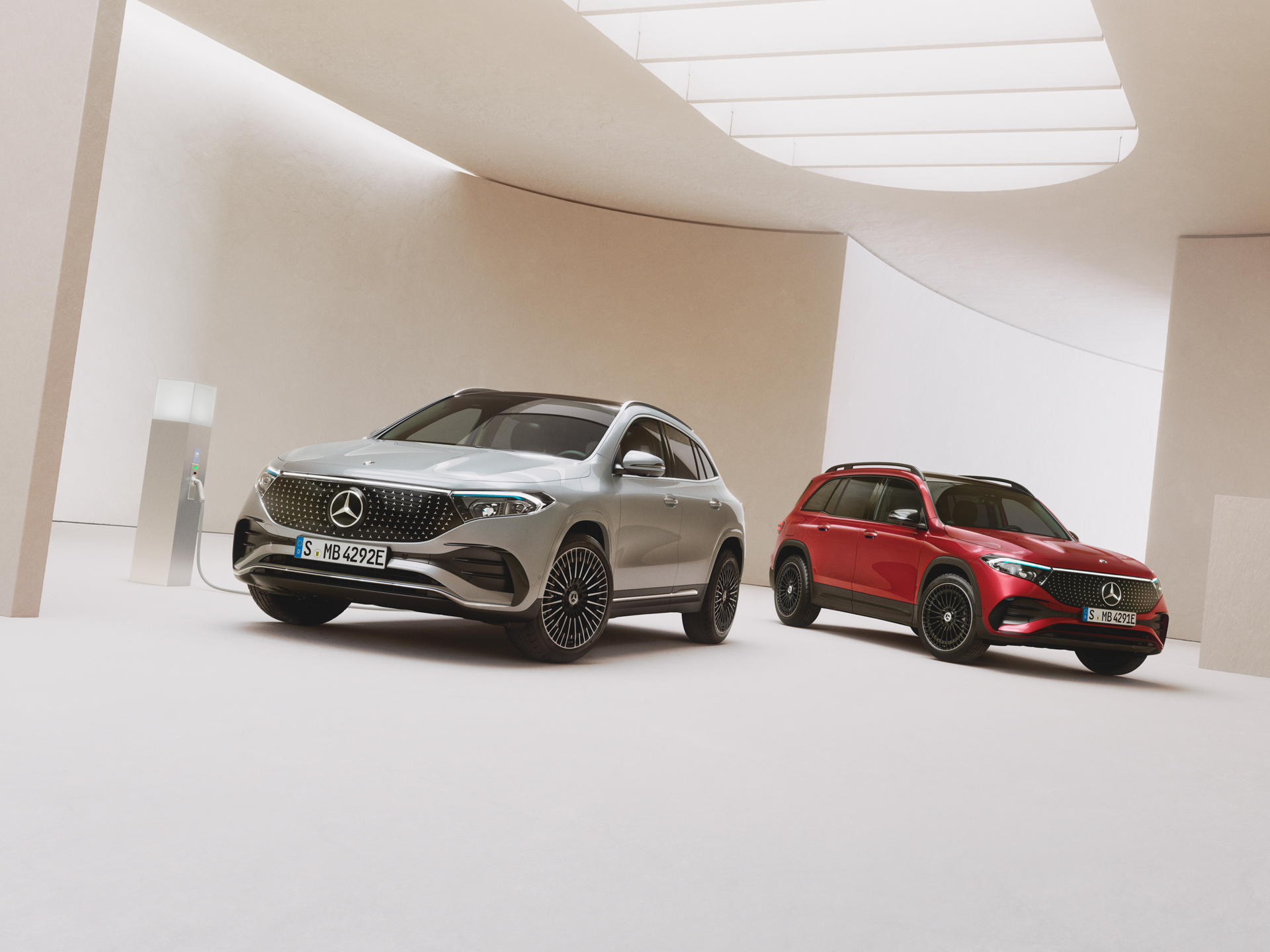 THG-Quoten | Elektroauto Förderungen | Mercedes-Benz. Ein roter Mercedes-Benz EQB und ein grauer Mercedes-Benz EQA stehen auf einem hellen Untergrund an einer Ladesäule.