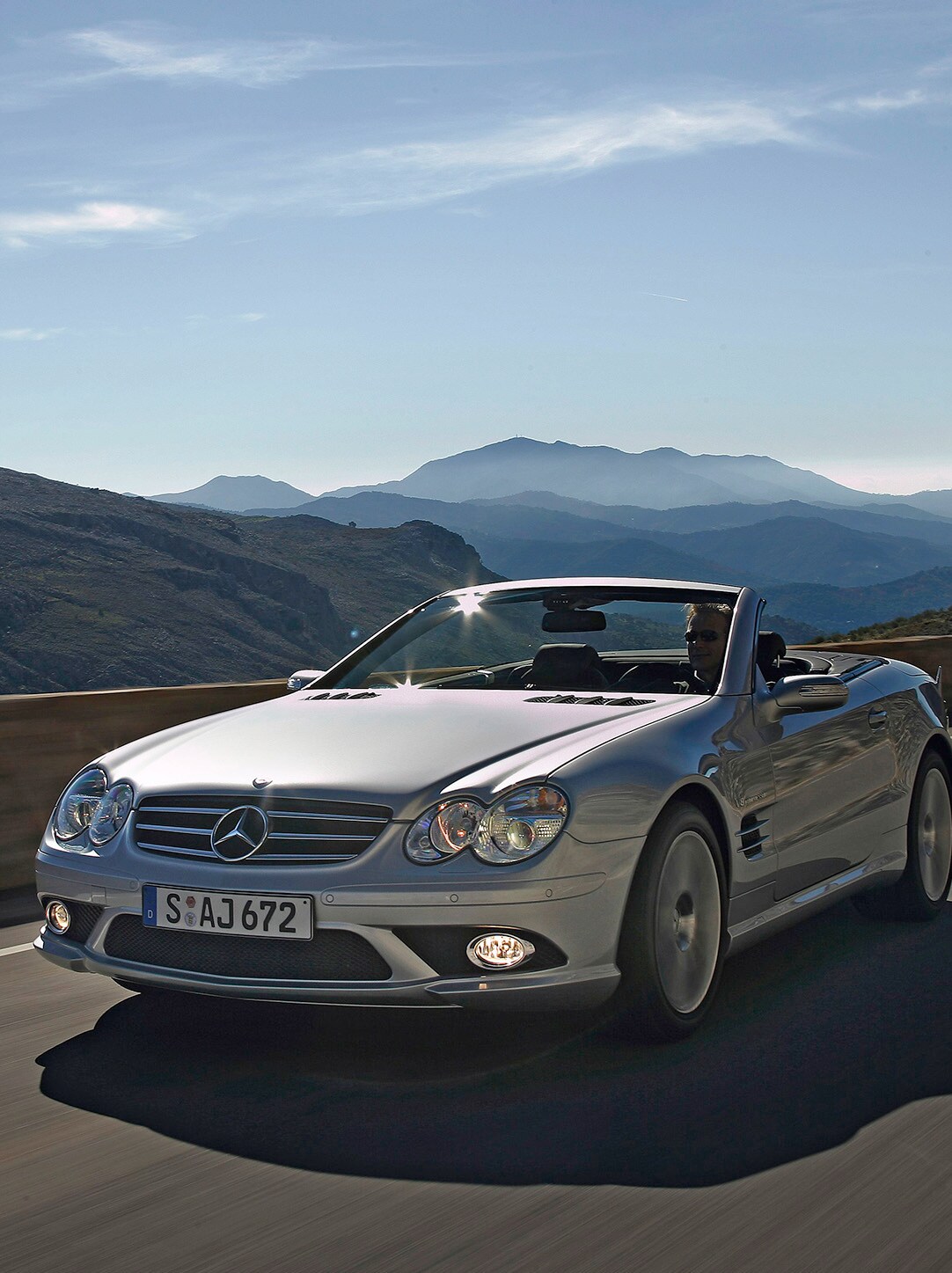 Ein silbernes Cabriolet von Mercedes-Benz fährt eine Straße mit Bergen entlang.