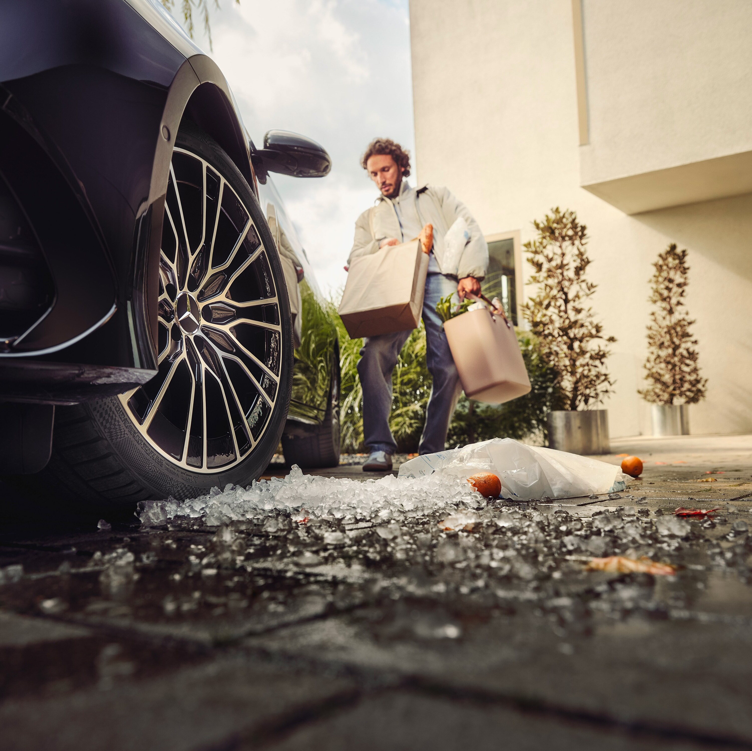 Ein Mann mit voll beladen mit Einkaufstüten geht zu seinem Mercedes-Benz, wobei einige Sachen schon auf dem Boden liegen.