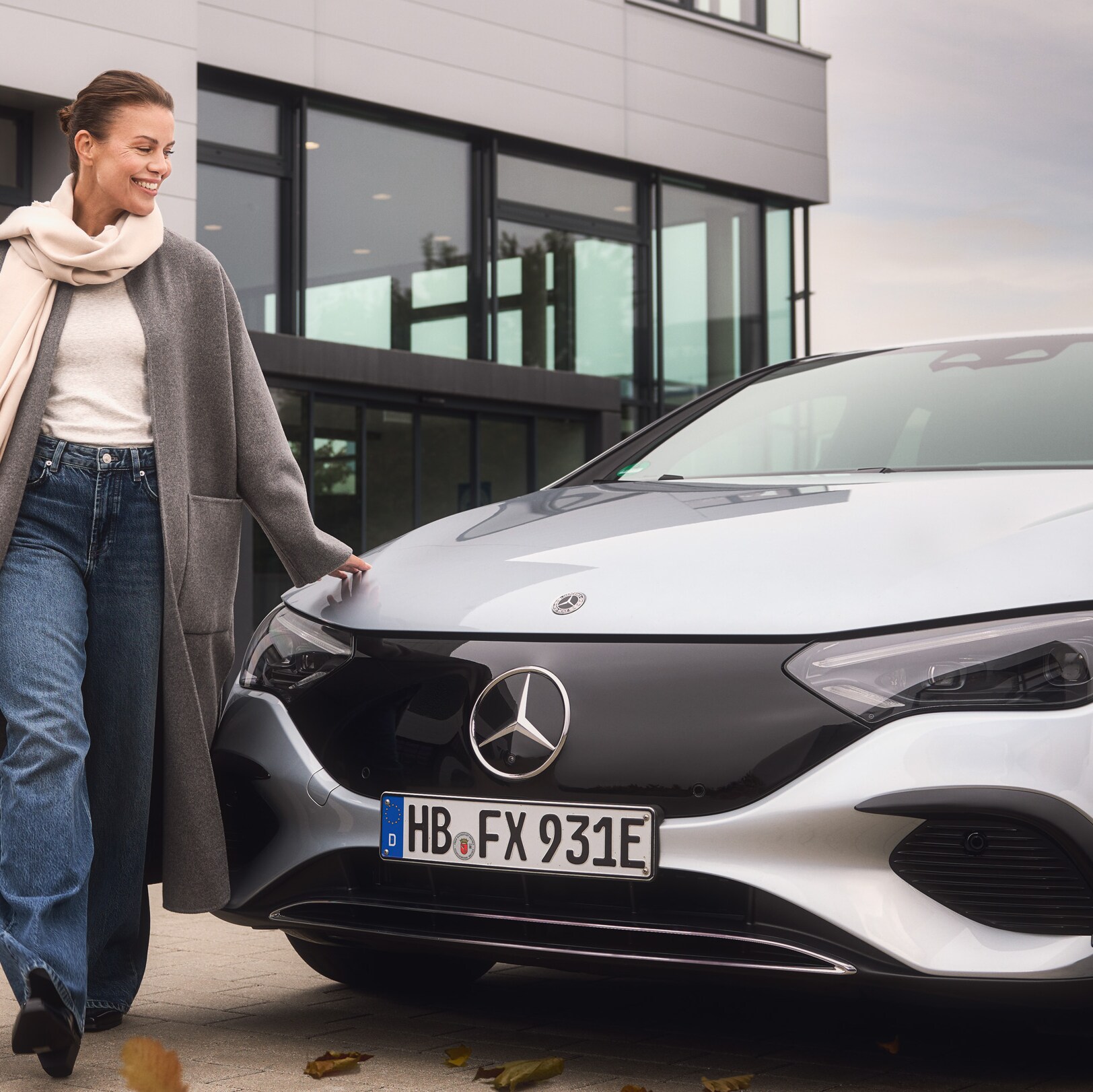 Eine Frau in einem herbstlichen Outfit steht vor einem silbernen Mercedes-Benz.