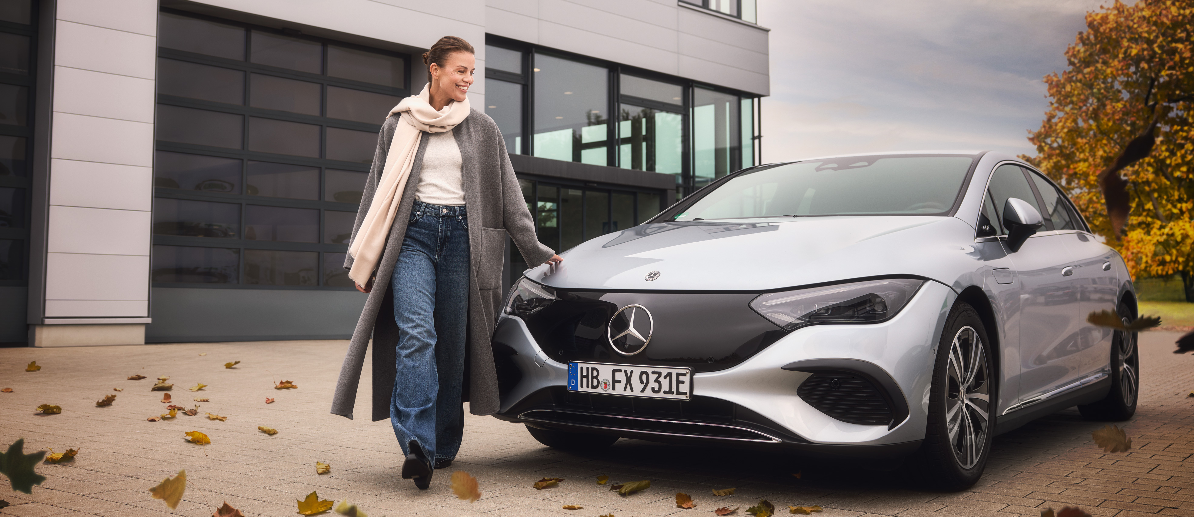 Herbstaktion | Mercedes-Benz Eine Frau in einem herbstlichen Outfit steht vor einem silbernen Mercedes-Benz.