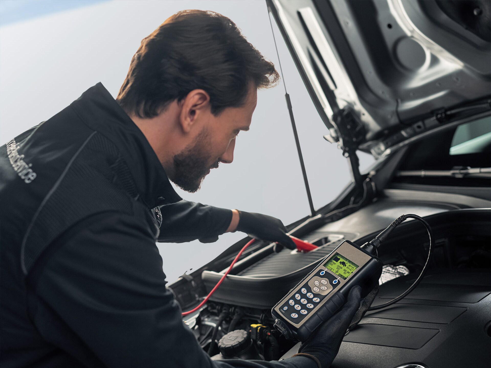 Ein junger Mercedes-Benz Mechaniker mit blauem Arbeitsanzug und Handschuhen prüft die Batterie eines Fahrzeugs.