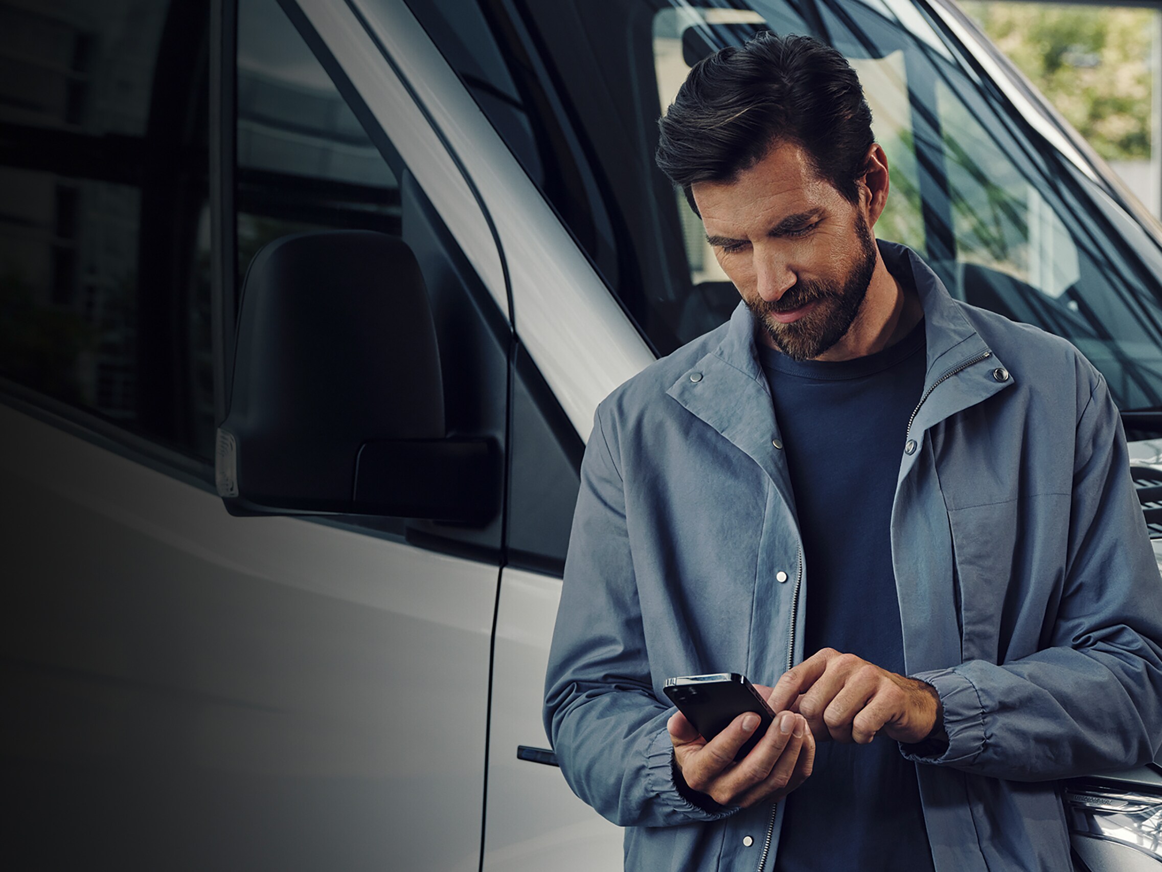 Ein Mann tippt auf seinem Handy und lächelt während er an einem Transporter lehnt.