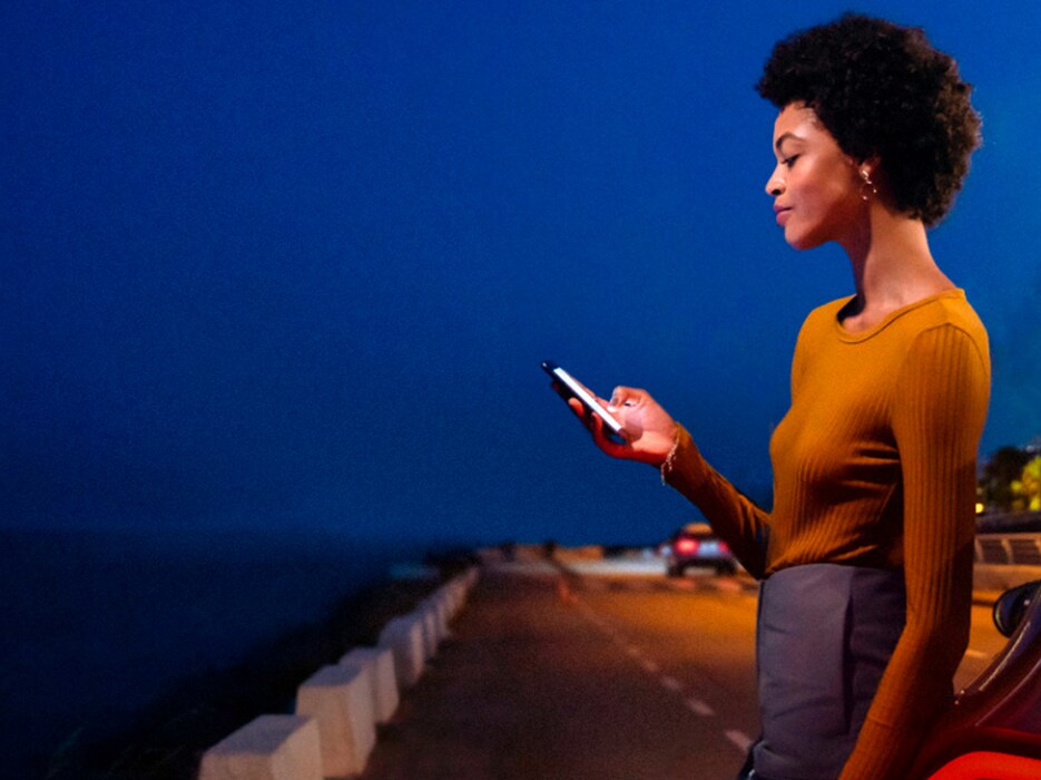 Eine Frau in gelbem Outfit schaut auf ihr Handy und steht dabei an einem Straßenrand.